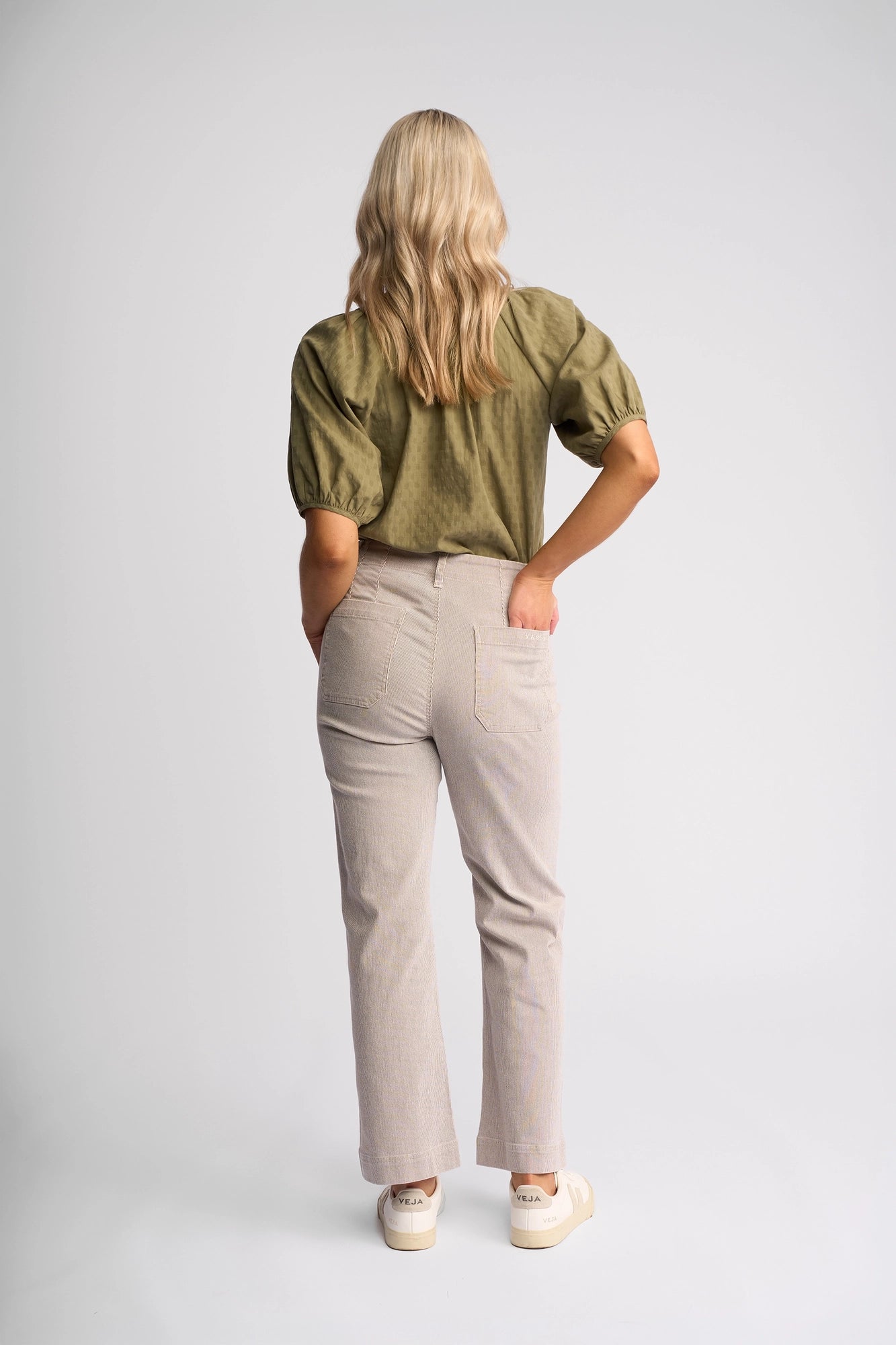 A woman with long blonde hair is standing with her back to the camera, wearing a green short-sleeve blouse, Vassalli Daily Jean in Ecru Stripe Denim, and white sneakers against a plain white background.