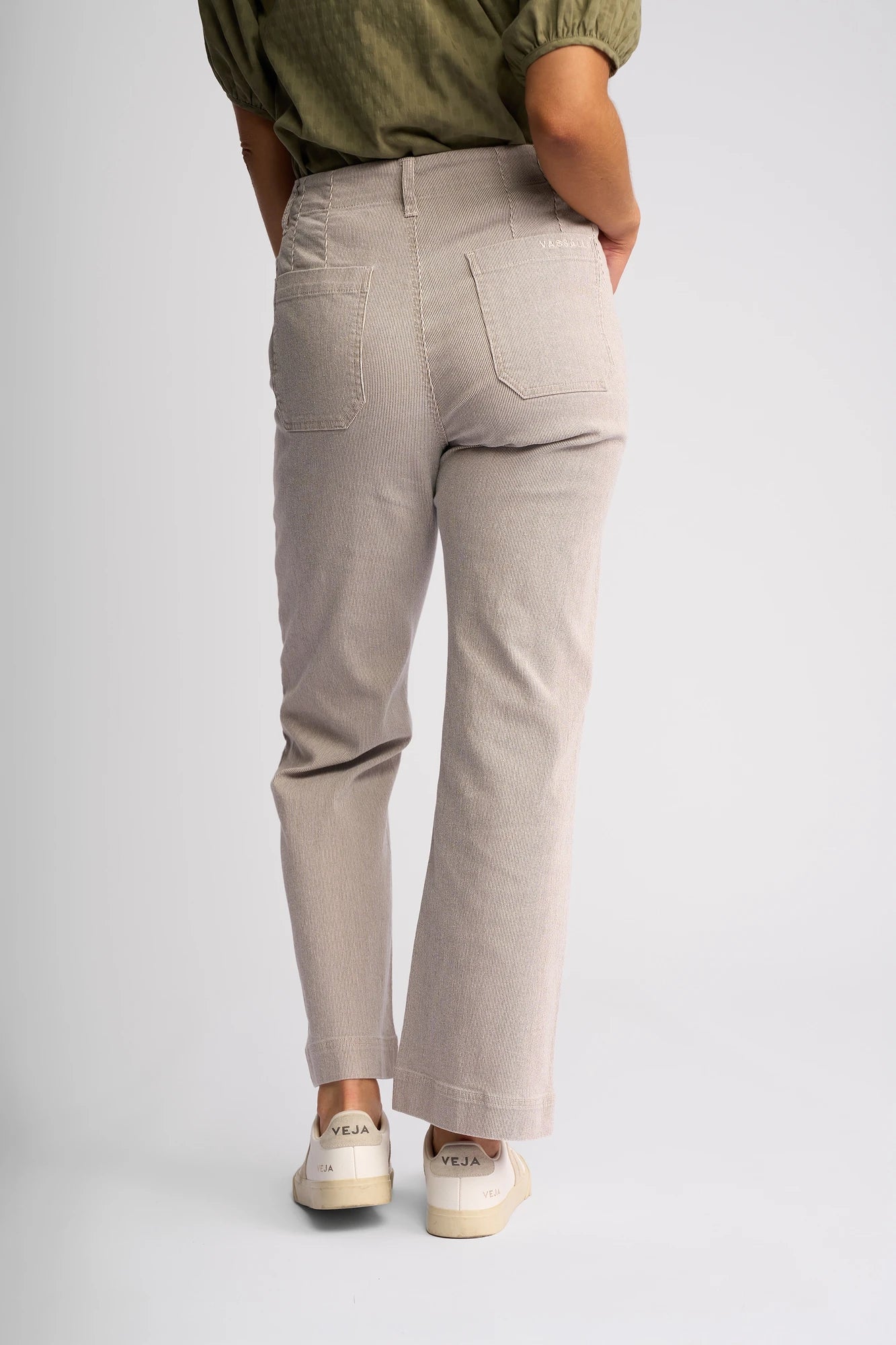 A person is standing with their back to the camera, wearing the Vassalli Daily Jean in Ecru Stripe Denim—light beige, straight-leg jeans—paired with a green top with rolled sleeves and white Veja sneakers, against a plain light background.