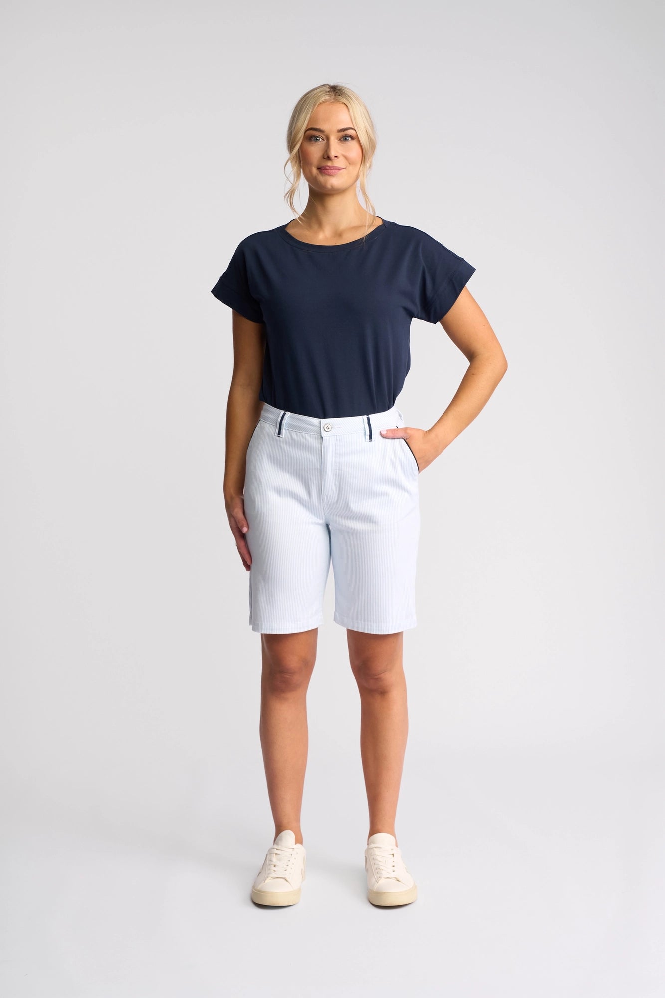 A woman stands facing forward, wearing a navy blue t-shirt and the Vassalli Orion Short in Blue Stripe Denim. She has one hand in her pocket and beige sneakers on her feet. The background is plain and light-colored.