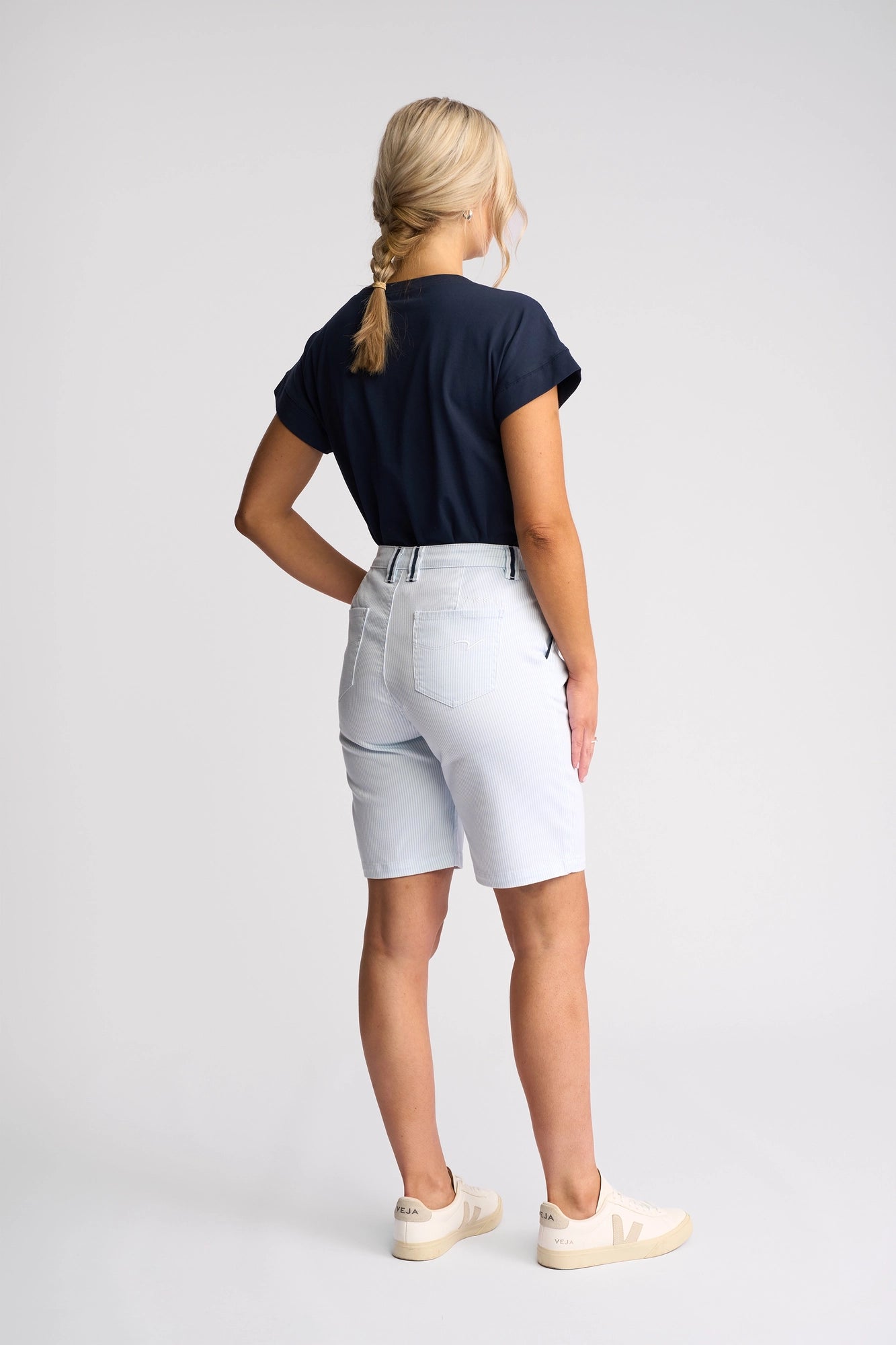 A woman with blonde hair in a braid, dressed in a navy t-shirt and Vassalli Orion Short - Blue Stripe Denim, paired with beige sneakers, stands facing away on a plain background.