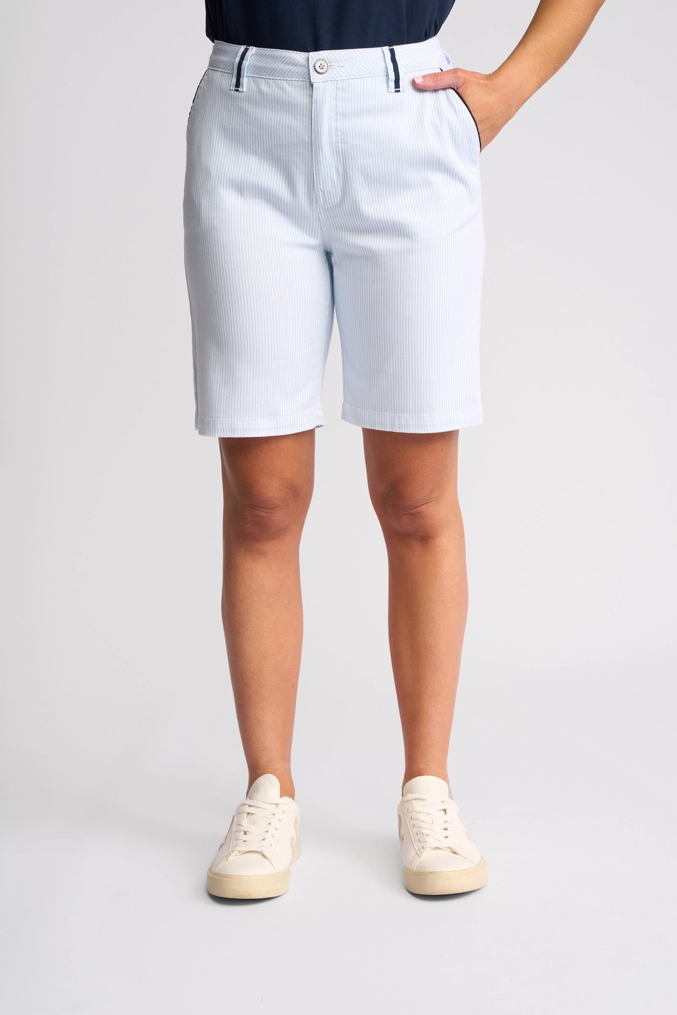 A person wearing the Vassalli Orion Short in Blue Stripe Denim, paired with a navy top and white sneakers, stands with one hand in their pocket against a plain background.