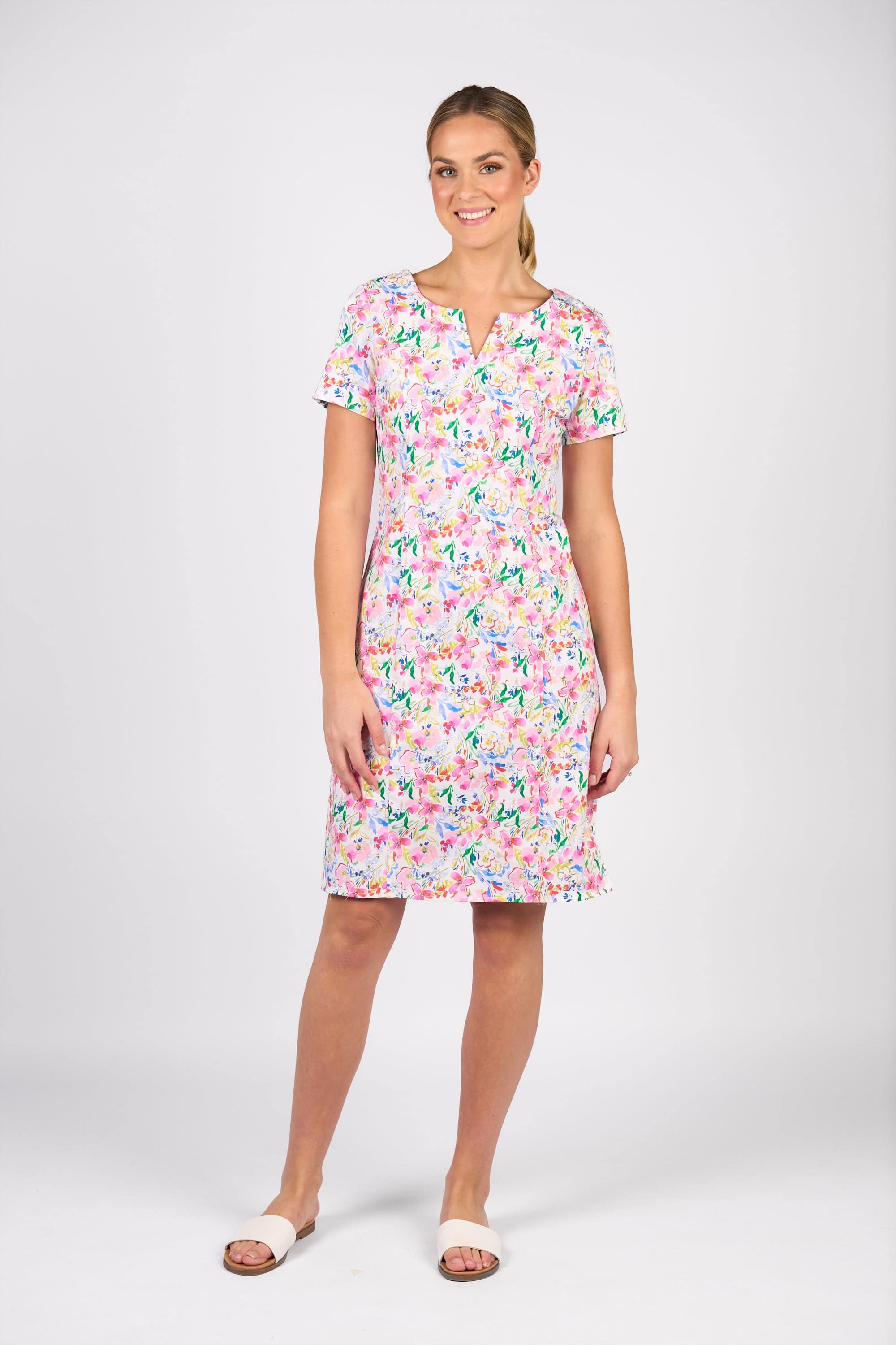 A woman smiles in the Vassalli Nadia Dress - Porto, a colorful floral, short-sleeved stretch cotton dress with a small V-neck notch. She pairs it with white slide sandals and wears her hair pulled back against a plain white background.