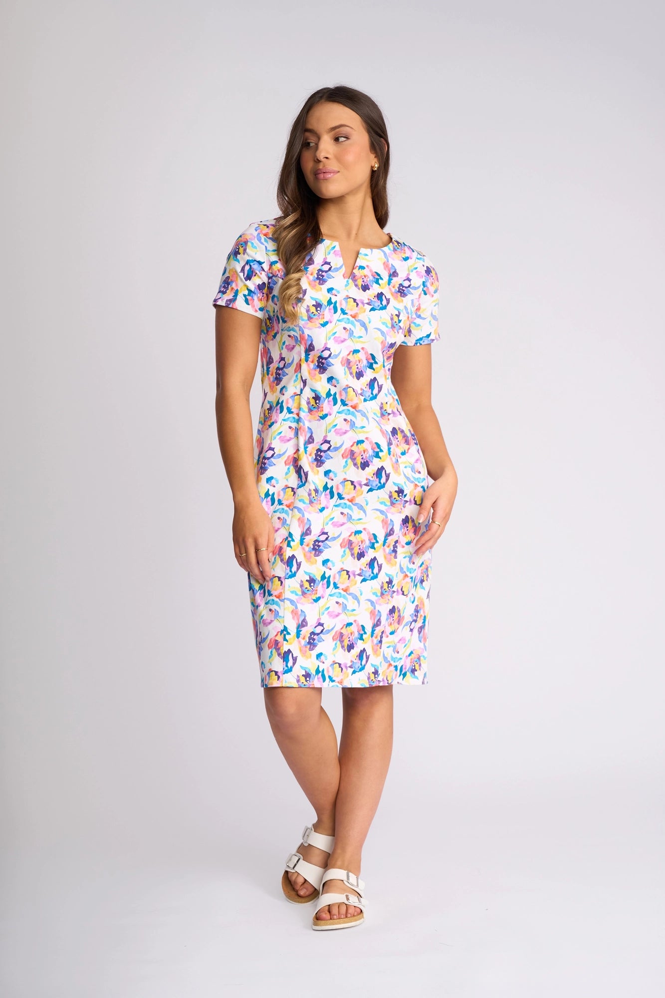 A woman stands in a white studio wearing the Vassalli Nadia Dress - Demi, a knee-length dress featuring a colorful floral print, crafted from stretch cotton with short sleeves. She pairs it with white sandals and looks slightly to the side, one hand in her pocket.