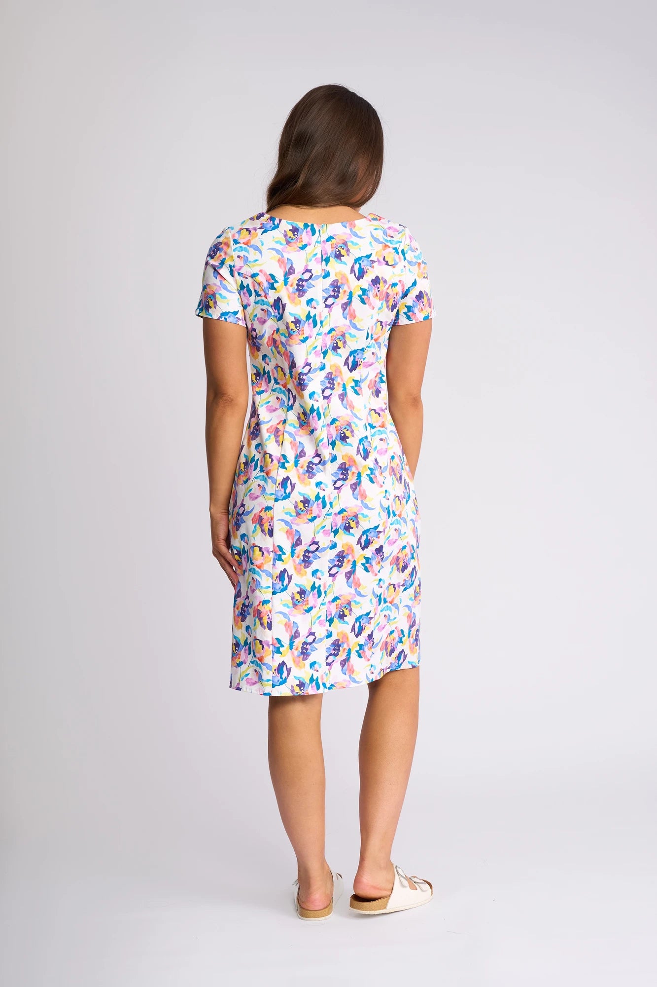A woman stands facing away, wearing the Vassalli Nadia Dress - Demi—a short-sleeved, knee-length floral print dress made from soft stretch cotton—paired with white slip-on shoes, against a plain light background.