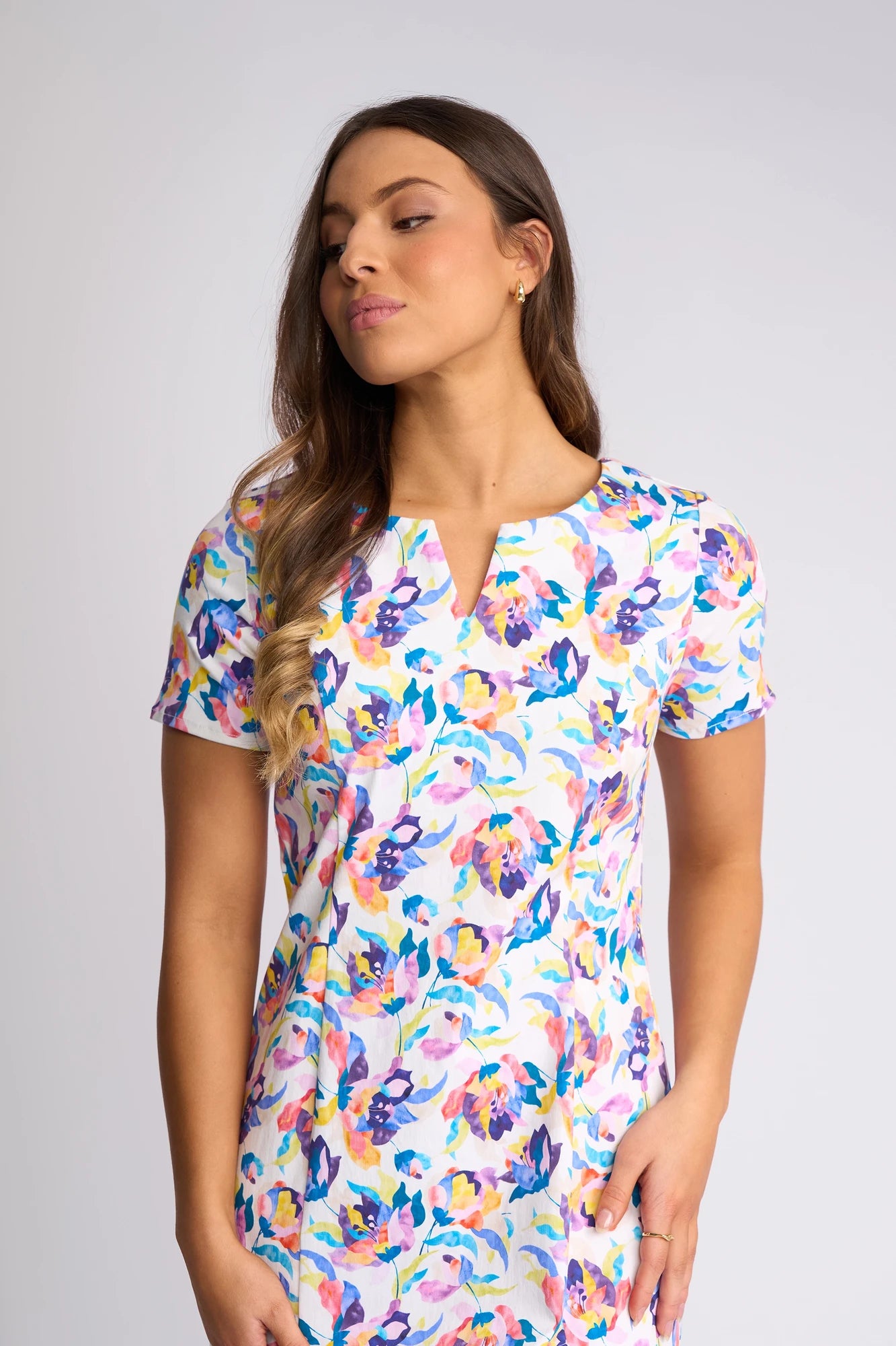 A woman wearing the Vassalli Nadia Dress - Demi, a short-sleeved floral print stretch cotton dress, stands against a plain light background, looking slightly to the side with one hand on her hip.