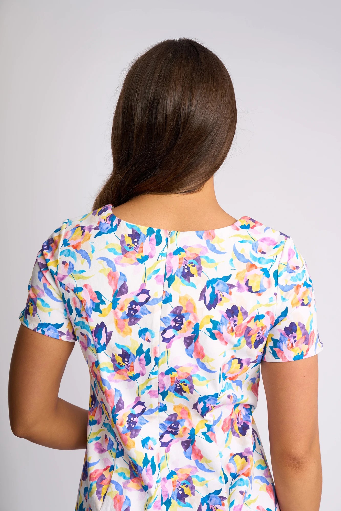 A person with long brown hair, seen from behind, wears the Vassalli Nadia Dress - Demi, a colorful floral print dress made from stretch cotton, against a plain light background.