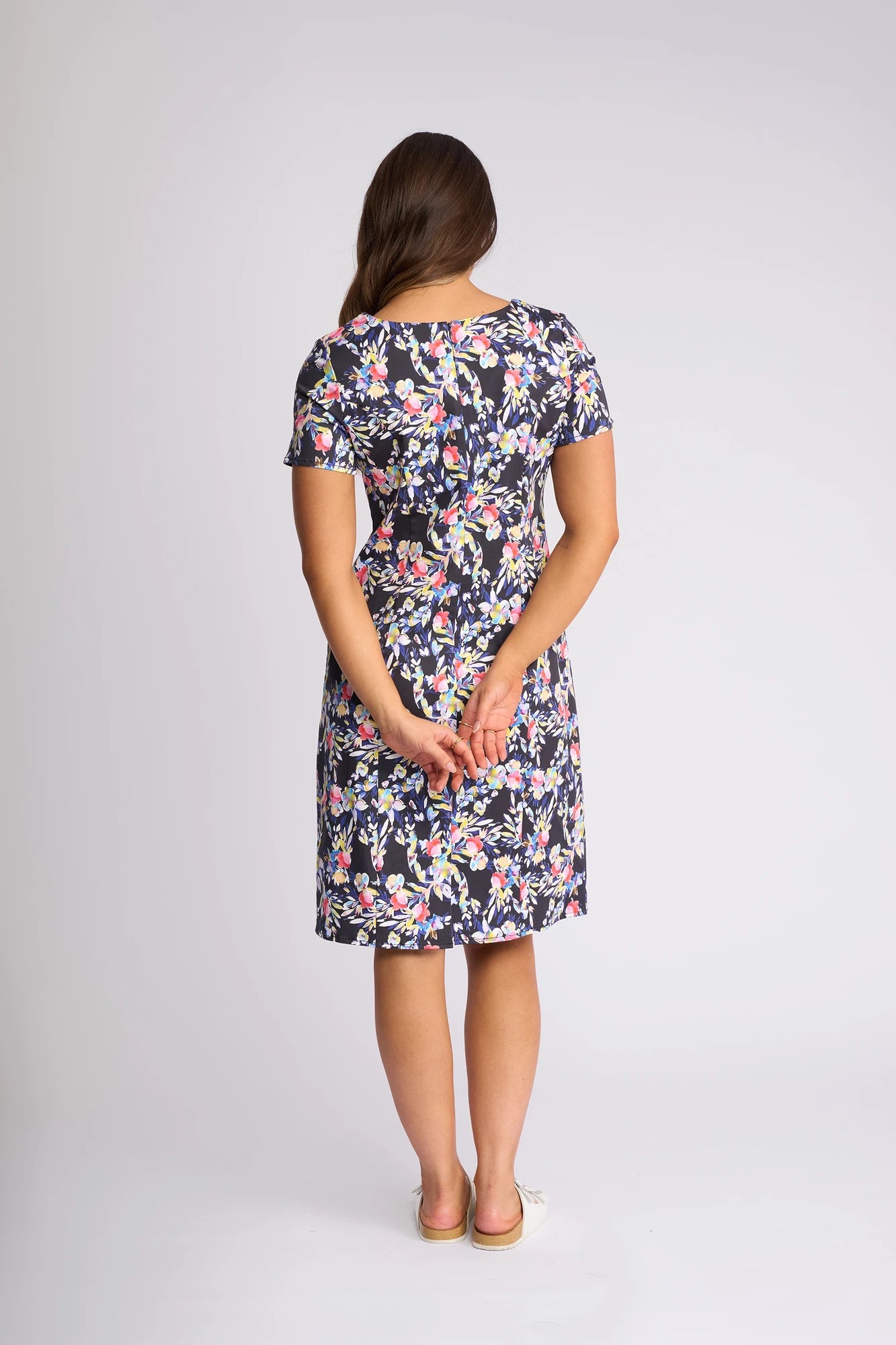 A woman stands facing away, hands clasped behind her back, wearing the Vassalli Nadia Dress - Lani—a short-sleeved, knee-length dress adorned with a dark floral pattern. Made from comfortable stretch cotton, the dress accentuates her long brown hair as she poses against a plain light-colored backdrop.