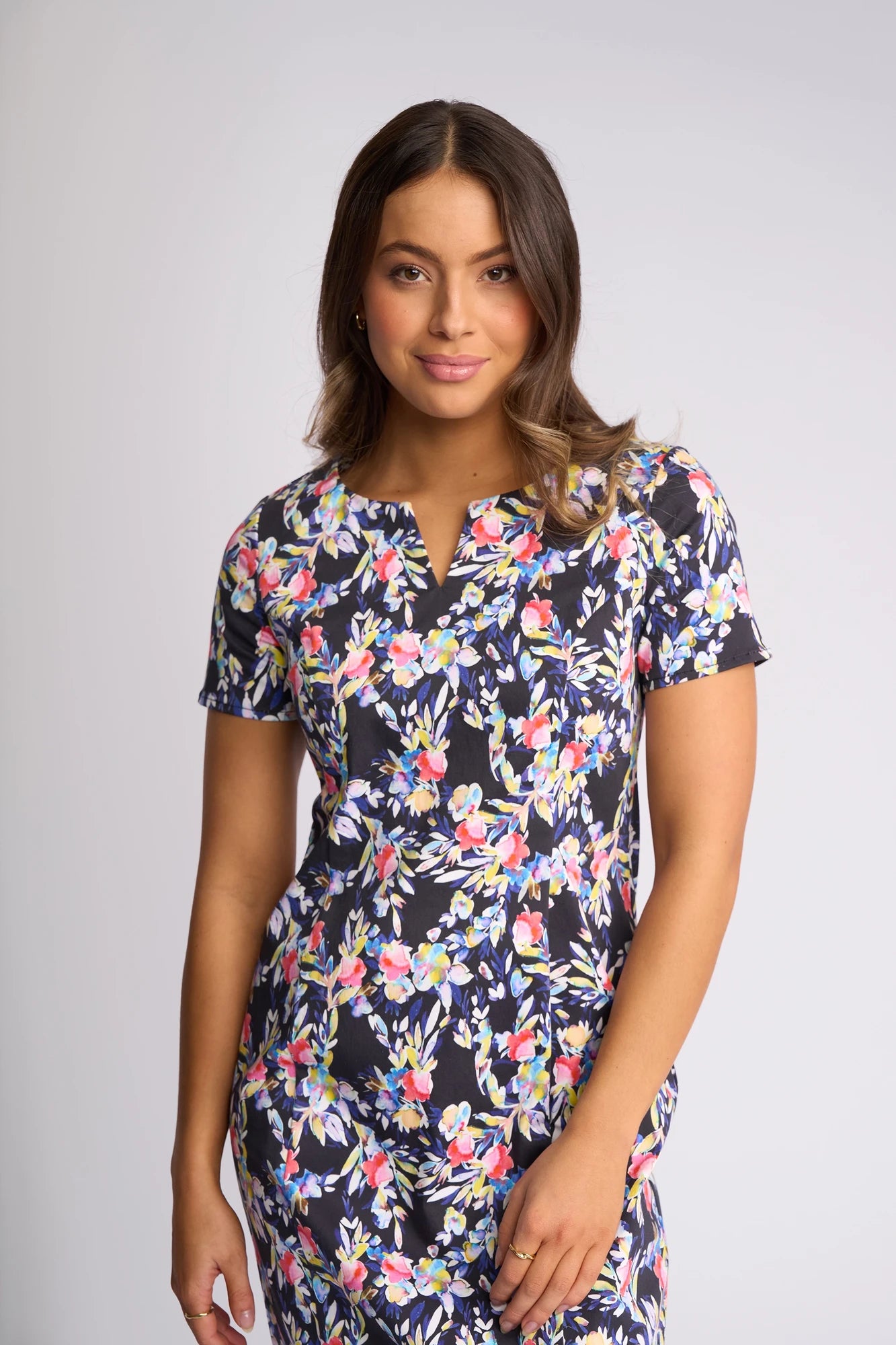 A woman with wavy brown hair, wearing a Vassalli Nadia Dress - Lani in a colorful floral print made from stretch cotton, stands against a light gray background and smiles softly at the camera.