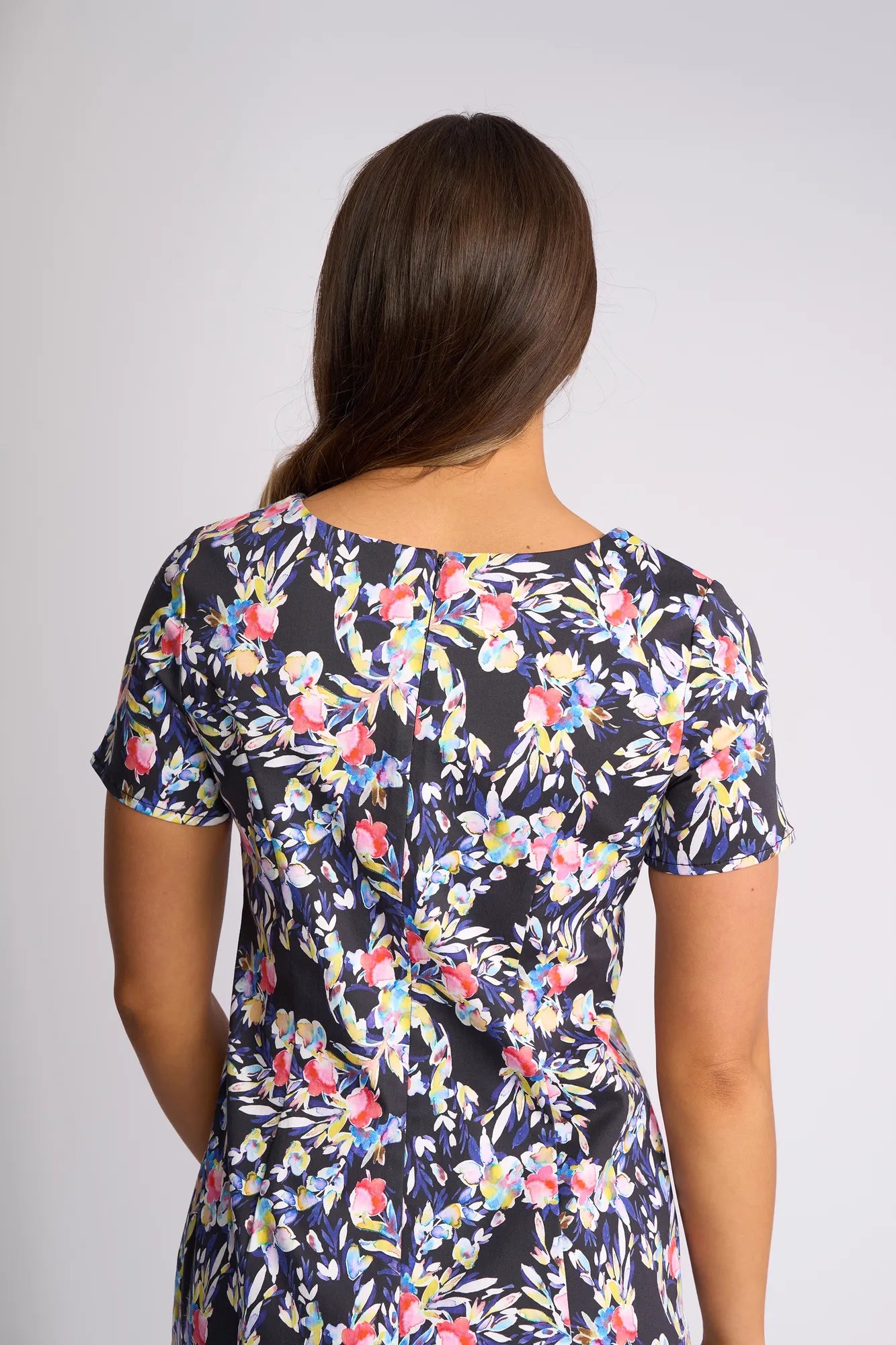 A person with long brown hair wearing the Vassalli Nadia Dress - Lani, a short-sleeved stretch cotton floral print dress, stands facing away from the camera against a plain, light background.