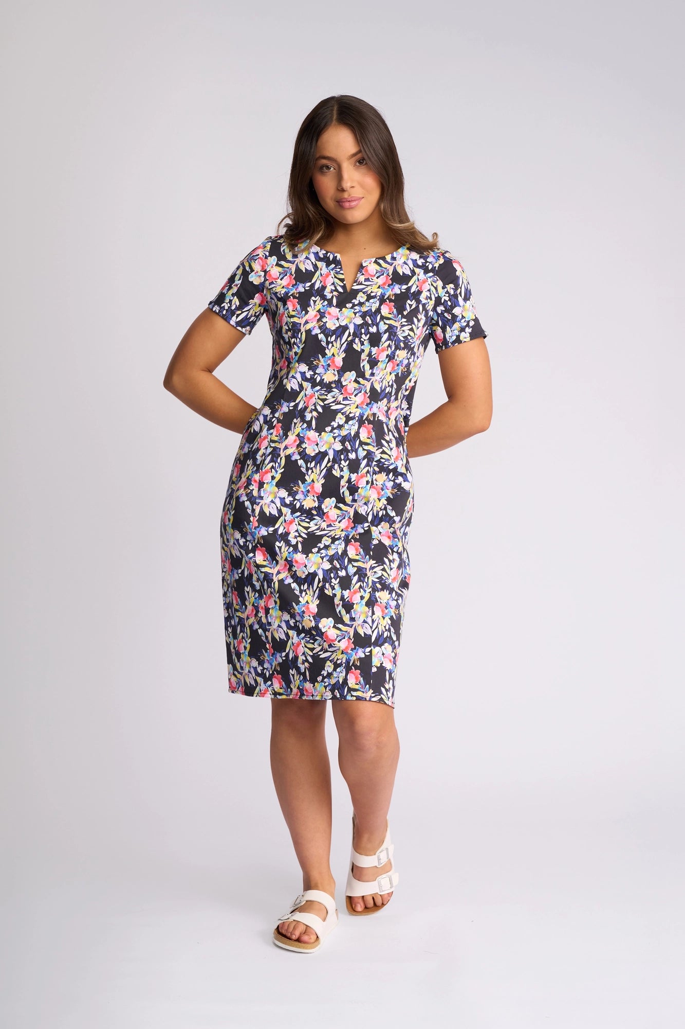 A woman stands against a plain light gray backdrop wearing the Vassalli Nadia Dress - Lani, a knee-length, short-sleeved floral print dress with a dark background made from stretch cotton. She has one hand behind her back and completes the look with white sandals.