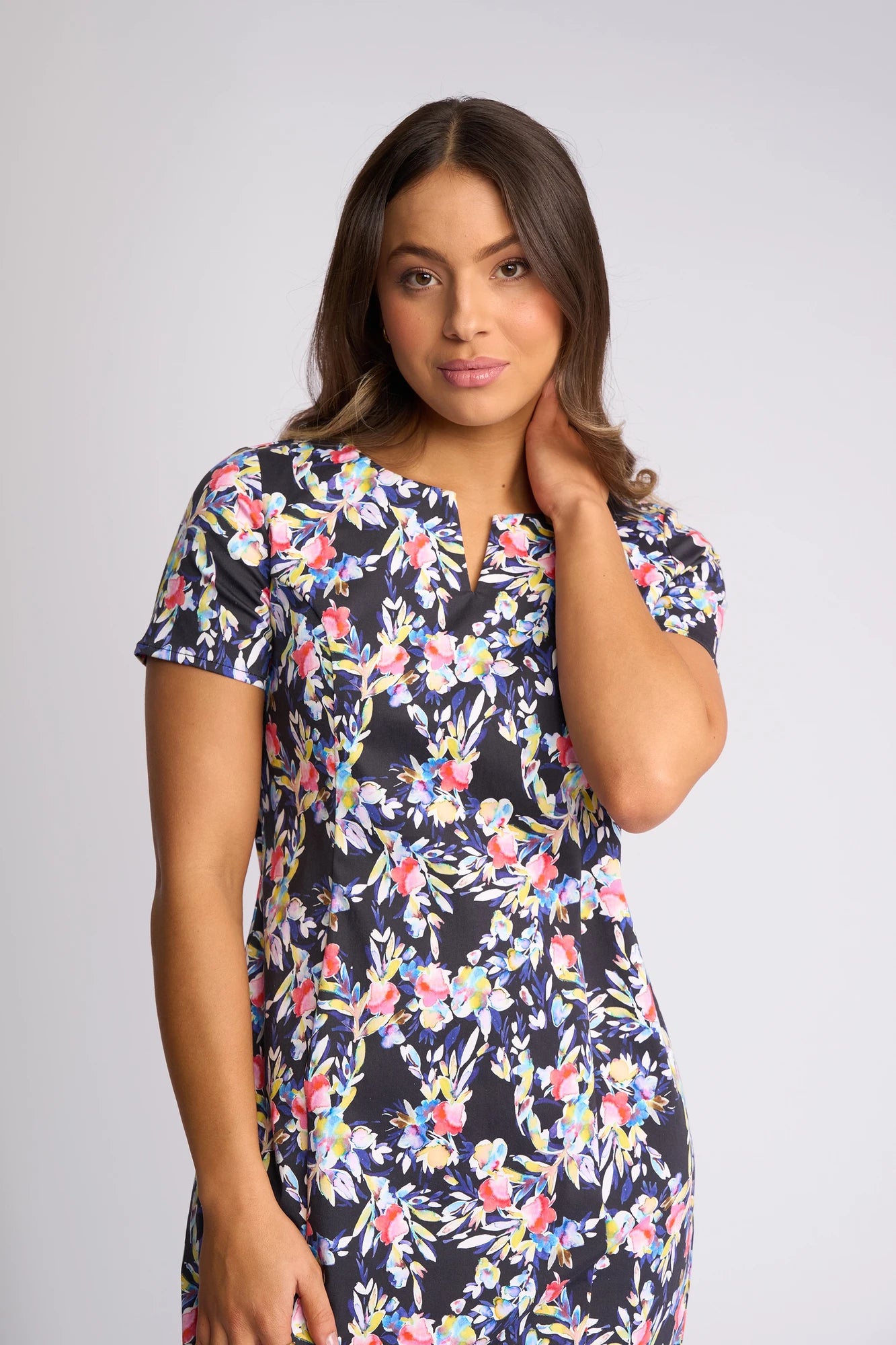 A woman with long brown hair wears the Vassalli Nadia Dress - Lani—a short-sleeved dress featuring a floral print with pink, blue, and white patterns—as she stands against a plain light background and touches her hair with one hand.