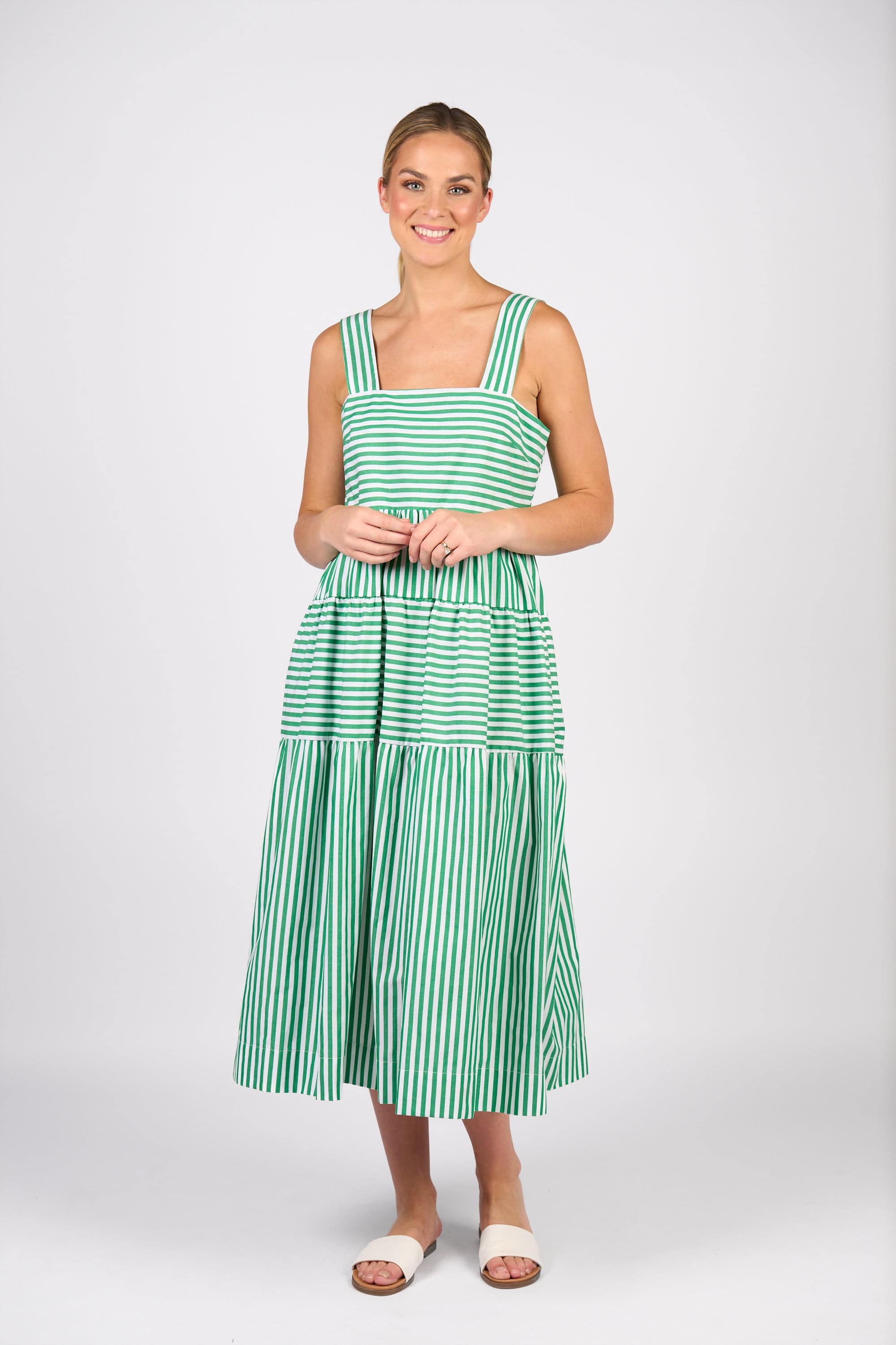 A woman with her light hair tied back wears the Vassalli Tiered Dress - Clover Stripe, featuring a sleeveless, tiered skirt. She pairs it with white slide sandals and stands against a plain white background, smiling.