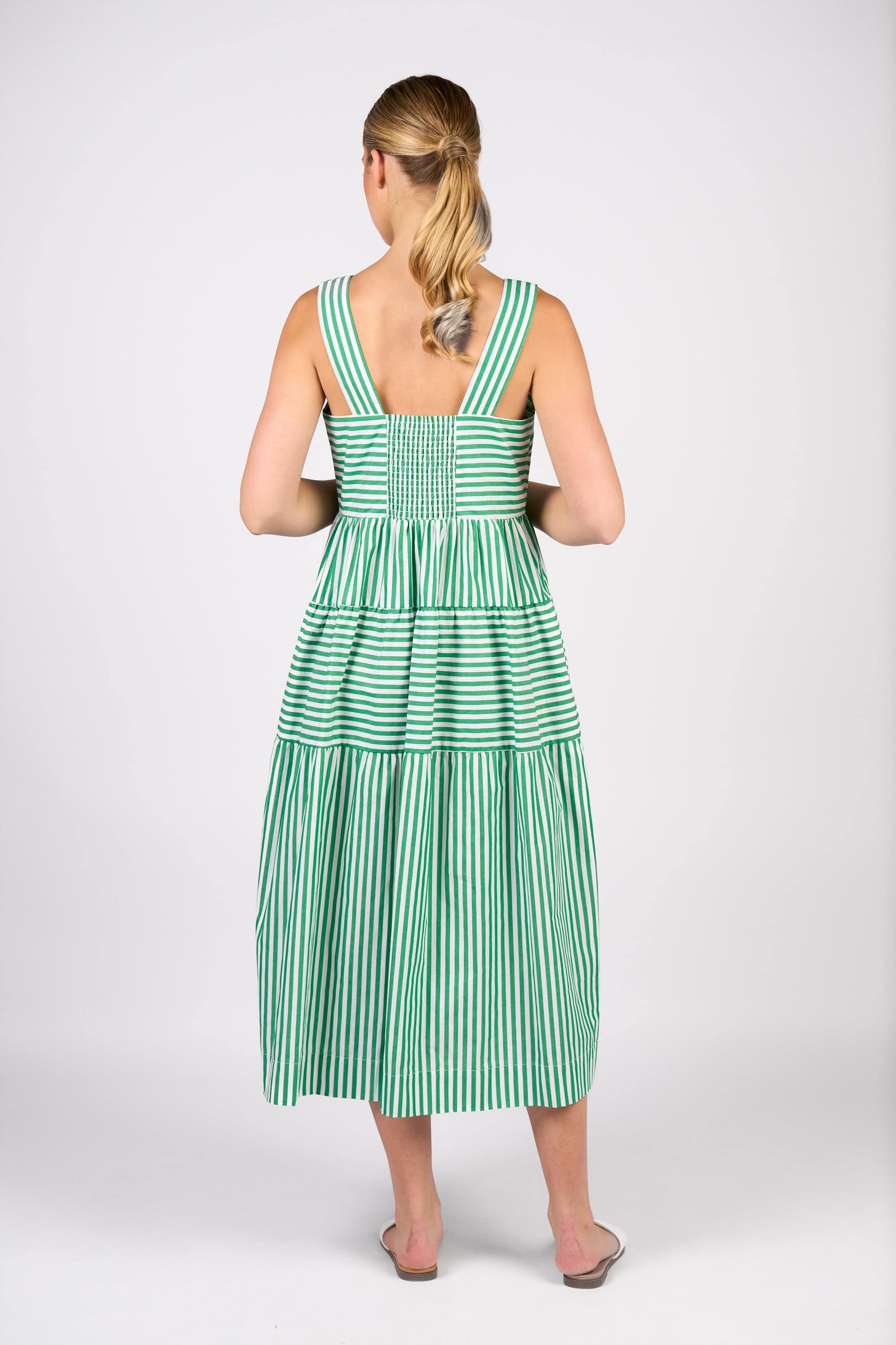 A woman with a low ponytail wears the Vassalli Tiered Dress - Clover Stripe, a sleeveless midi with wide straps, and white flats. She stands facing away against a plain white background.