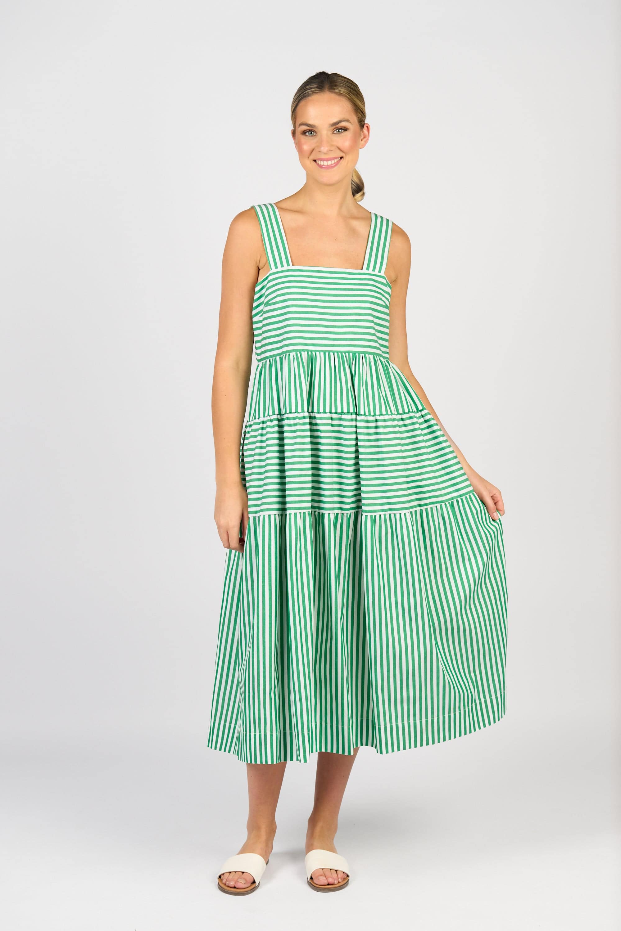 A woman wears the Vassalli Tiered Dress - Clover Stripe, smiling as she holds one side of the sleeveless, wide-strap dress and stands against a plain light background. She pairs it with white slide sandals.