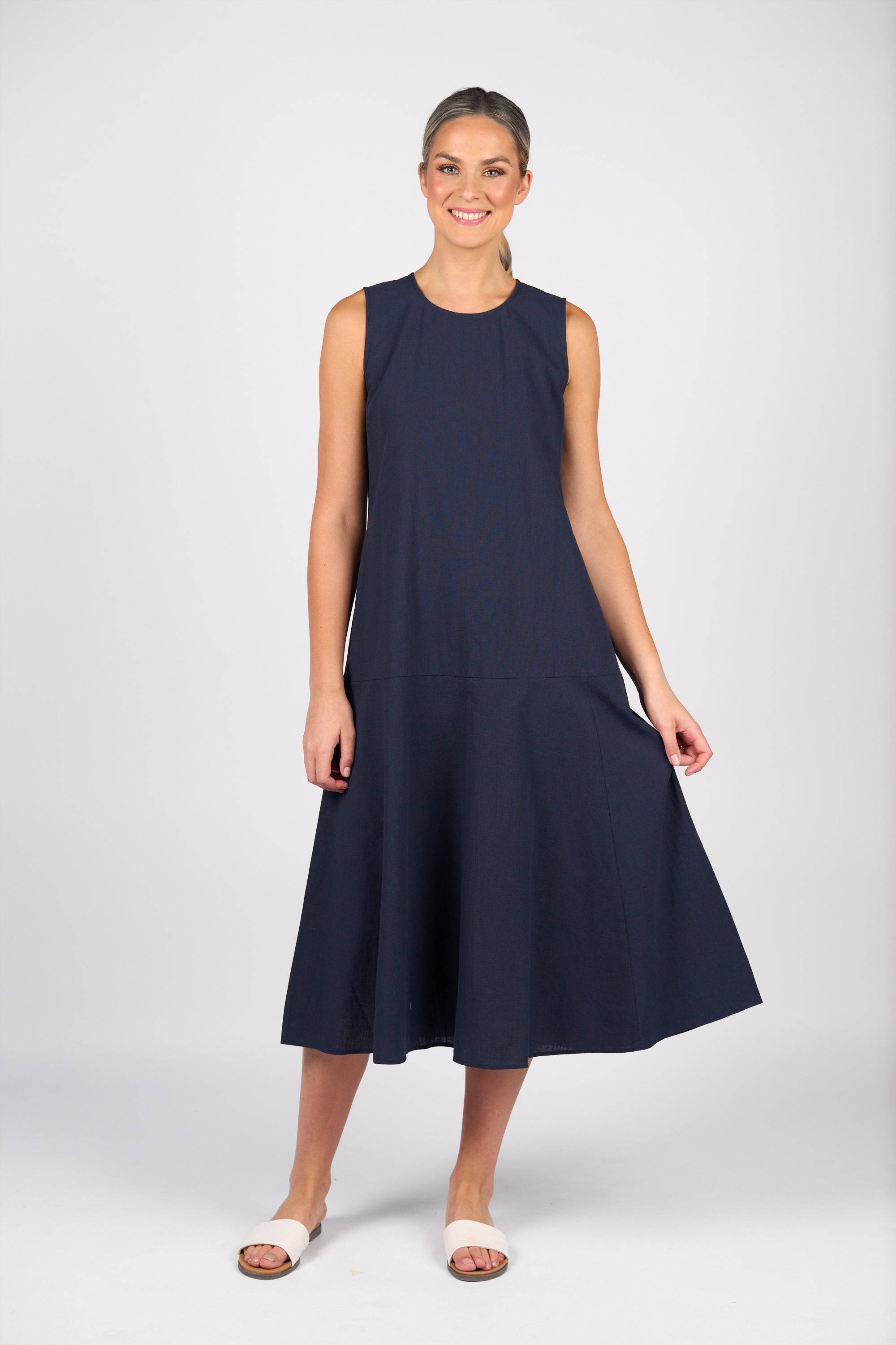 A woman with light skin and tied-back hair smiles in a Vassalli Sleeveless Dress - Ink, a 100% cotton navy fit-and-flare style with a flared skirt, paired with light sandals against a plain background.