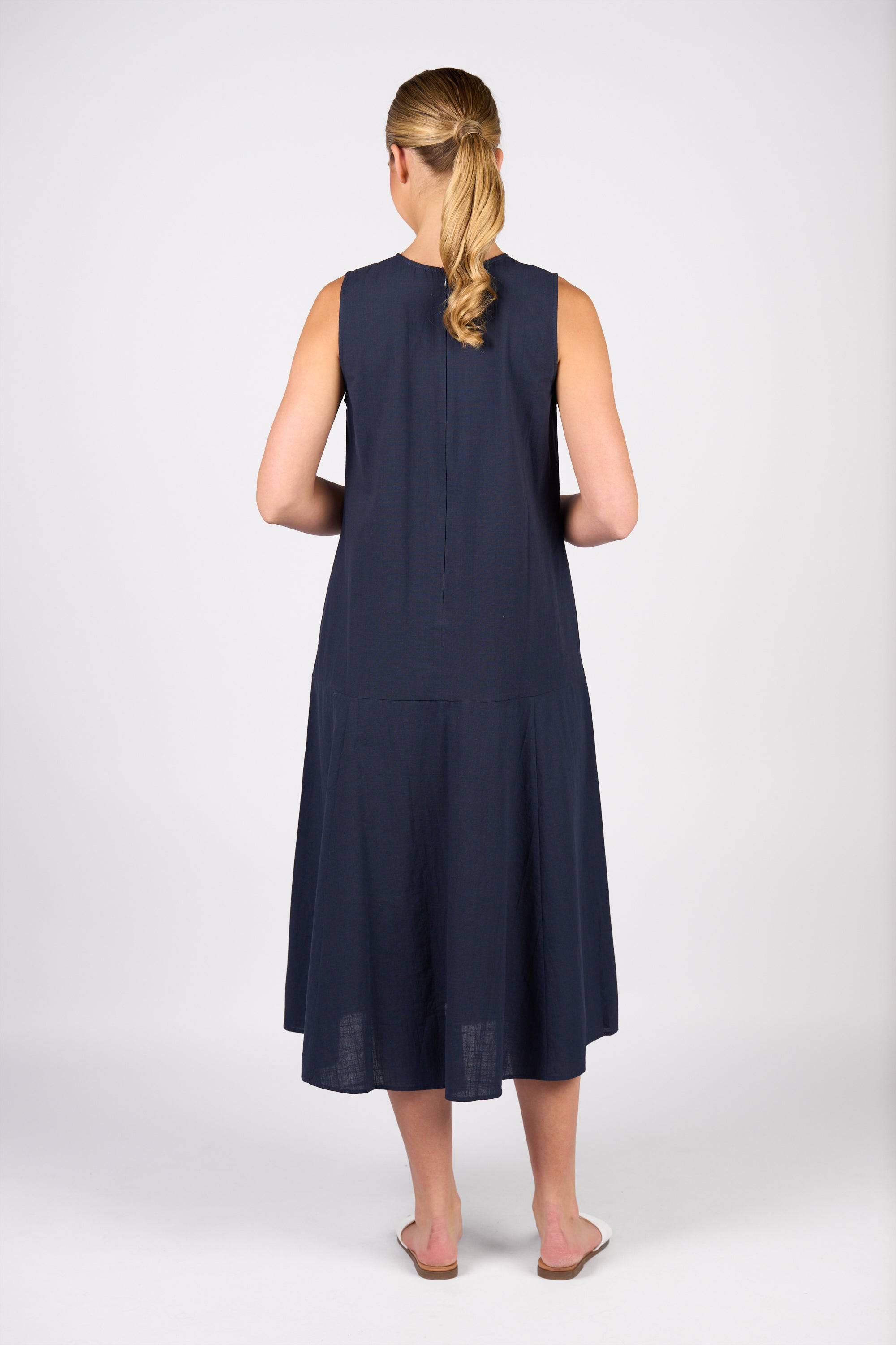 A woman with blonde hair in a low ponytail stands facing away, wearing the Vassalli Sleeveless Dress - Ink, made from 100% cotton, paired with white flat shoes against a plain white background.