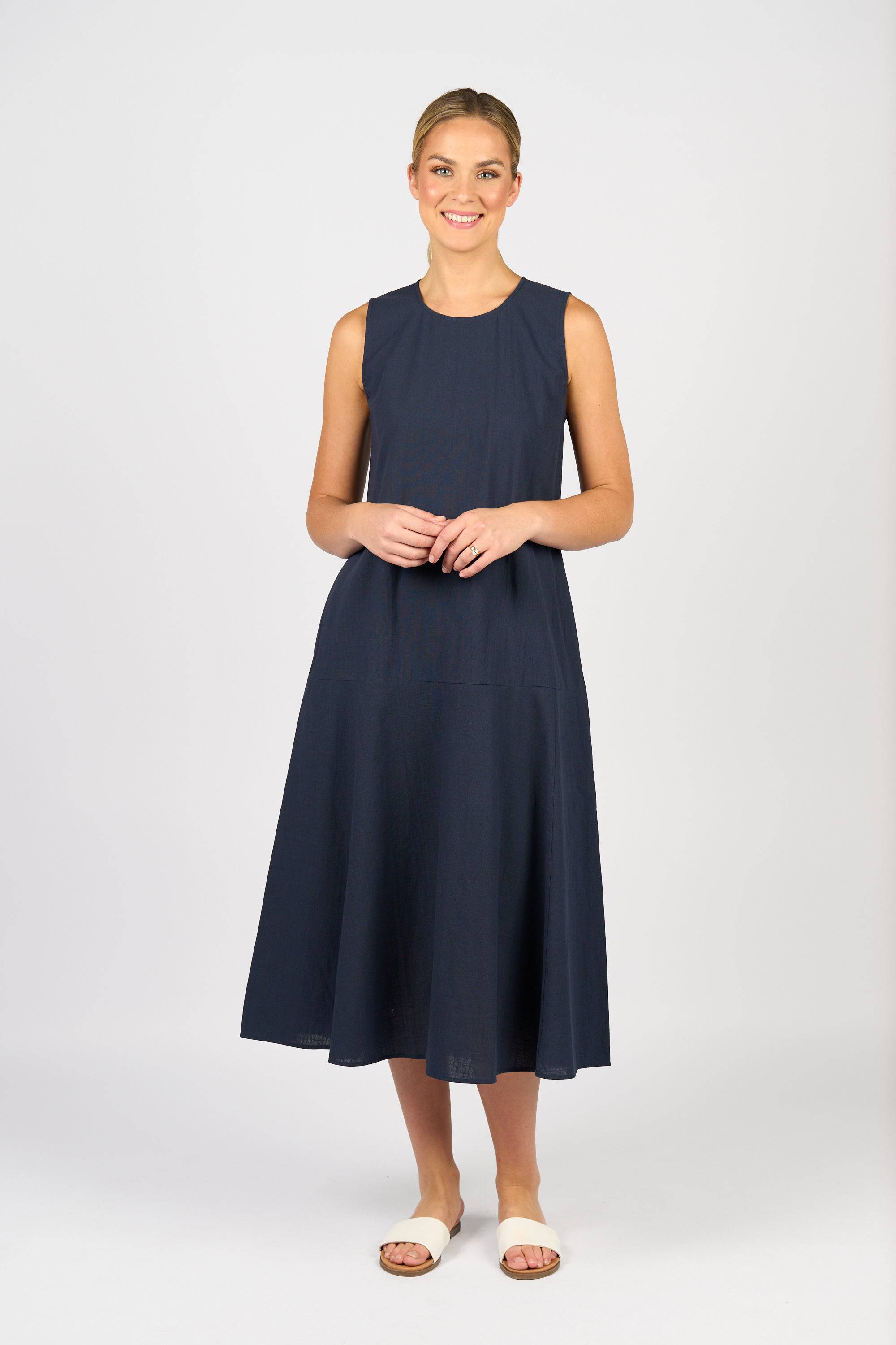 A woman smiles against a plain light background, wearing the Vassalli Sleeveless Dress - Ink and light-colored slide sandals.