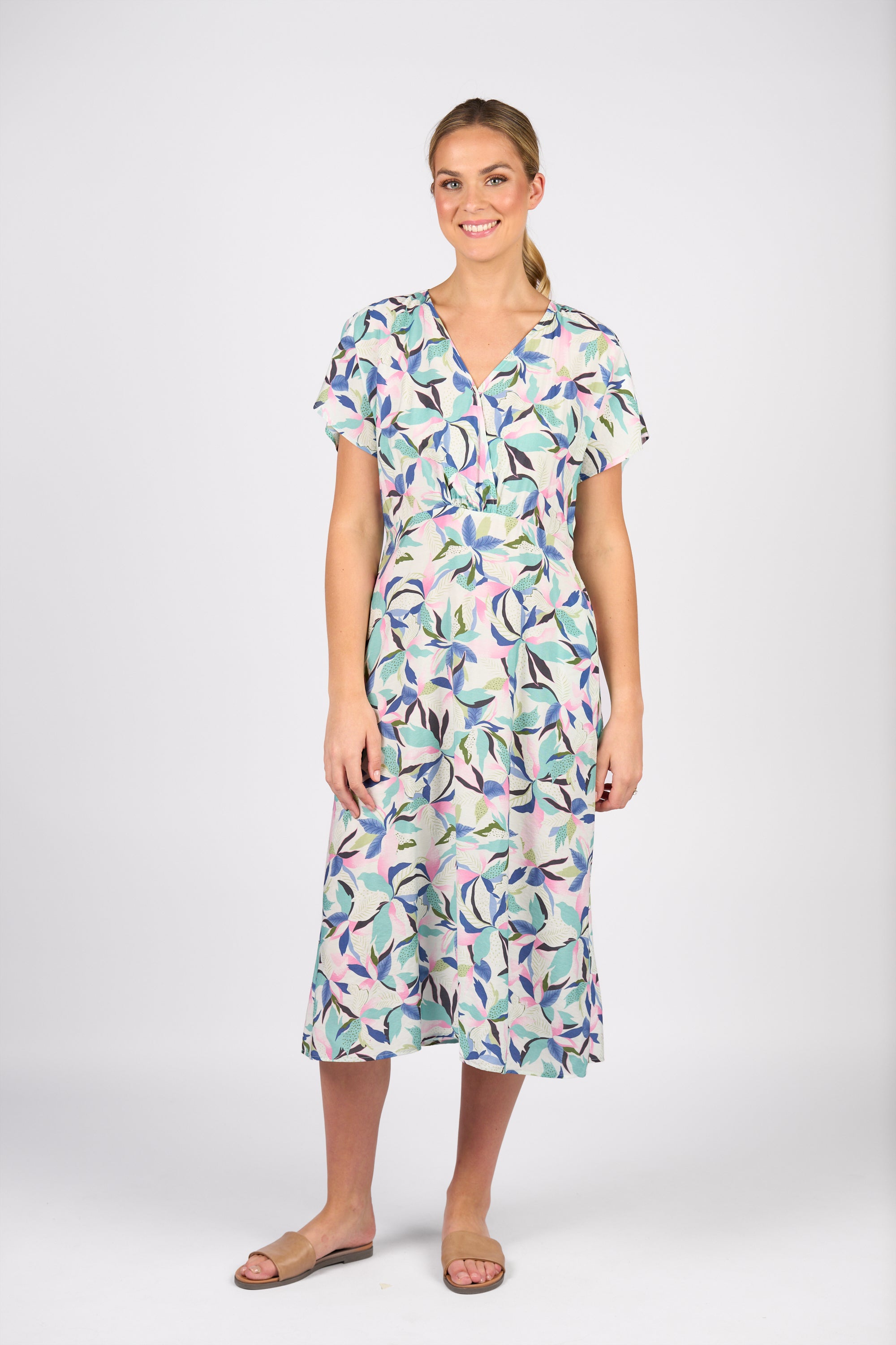 A woman smiles in the Vassalli Drop Shoulder Dress - Soleil, a light-toned, short-sleeved midi with a bold abstract leaf print and wrap bodice. She pairs it with tan sandals, hair tied back, posing against a plain white background.