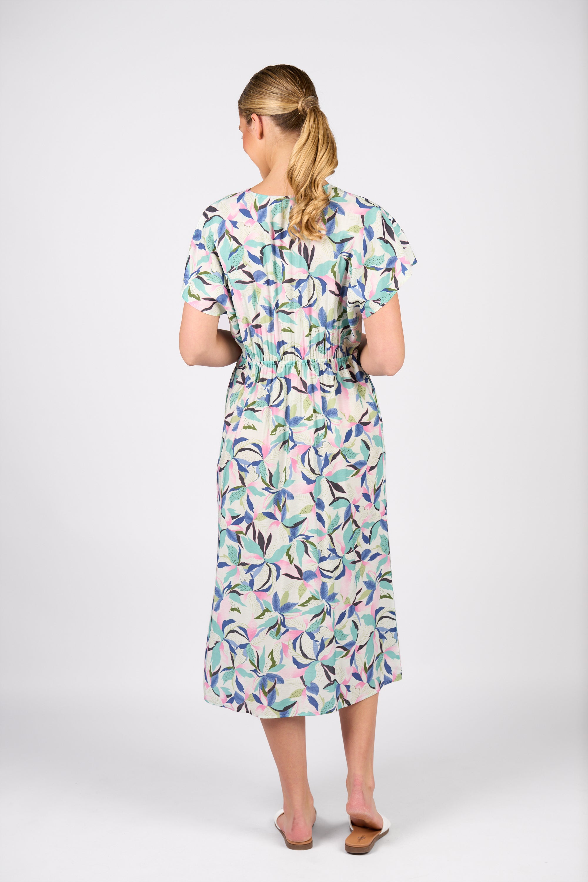 A woman with blonde hair in a low ponytail wears the Vassalli Drop Shoulder Dress - Soleil, showcasing its bold, colorful abstract leaf print and midi length. She faces away and pairs the dress with flat, tan sandals.