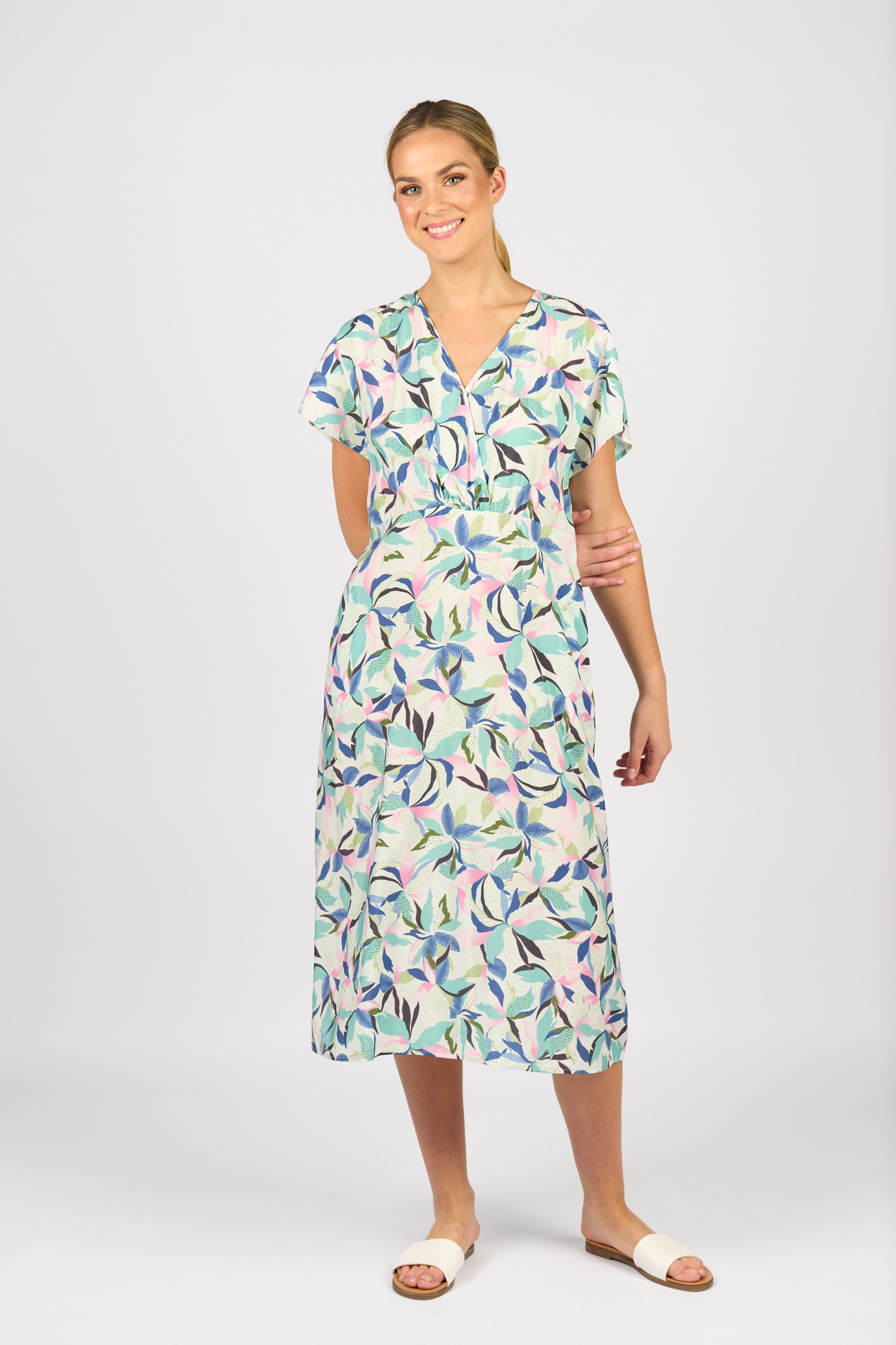 A woman smiles against a white background, wearing the Vassalli Drop Shoulder Dress - Soleil, a light midi dress with pastel leaf print and short sleeves, paired with white open-toe sandals. Her hair is pulled back.