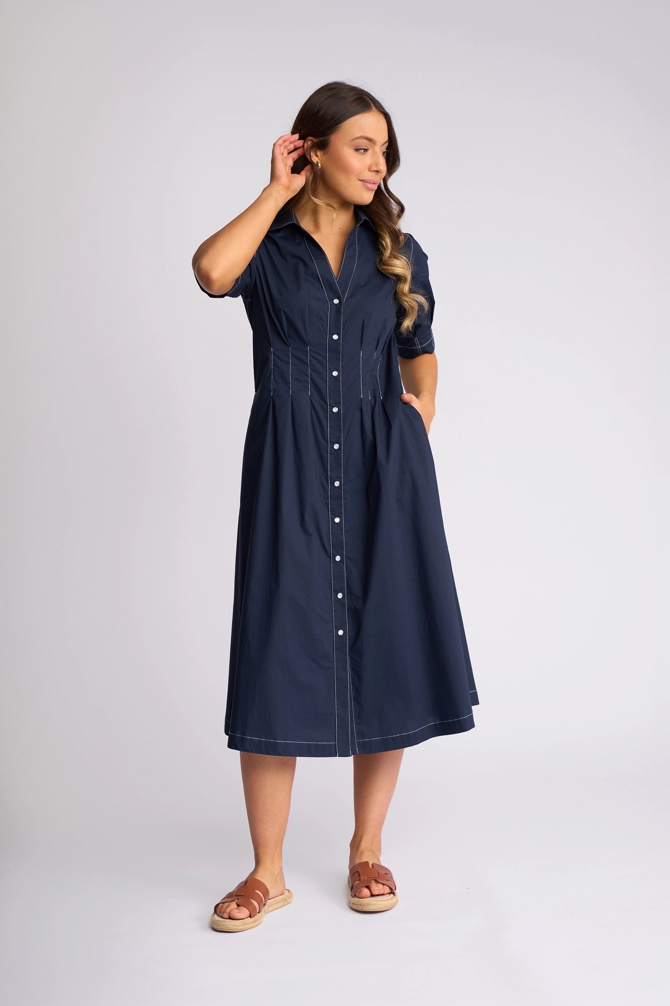 A woman wearing the Vassalli Addie Dress - Ink, a navy blue short-sleeve, button-down cotton dress with pockets, stands against a white background. She wears tan sandals, with one hand touching her hair and the other in her pocket. The feminine silhouette of the dress from Vassalli effortlessly flatters her figure.