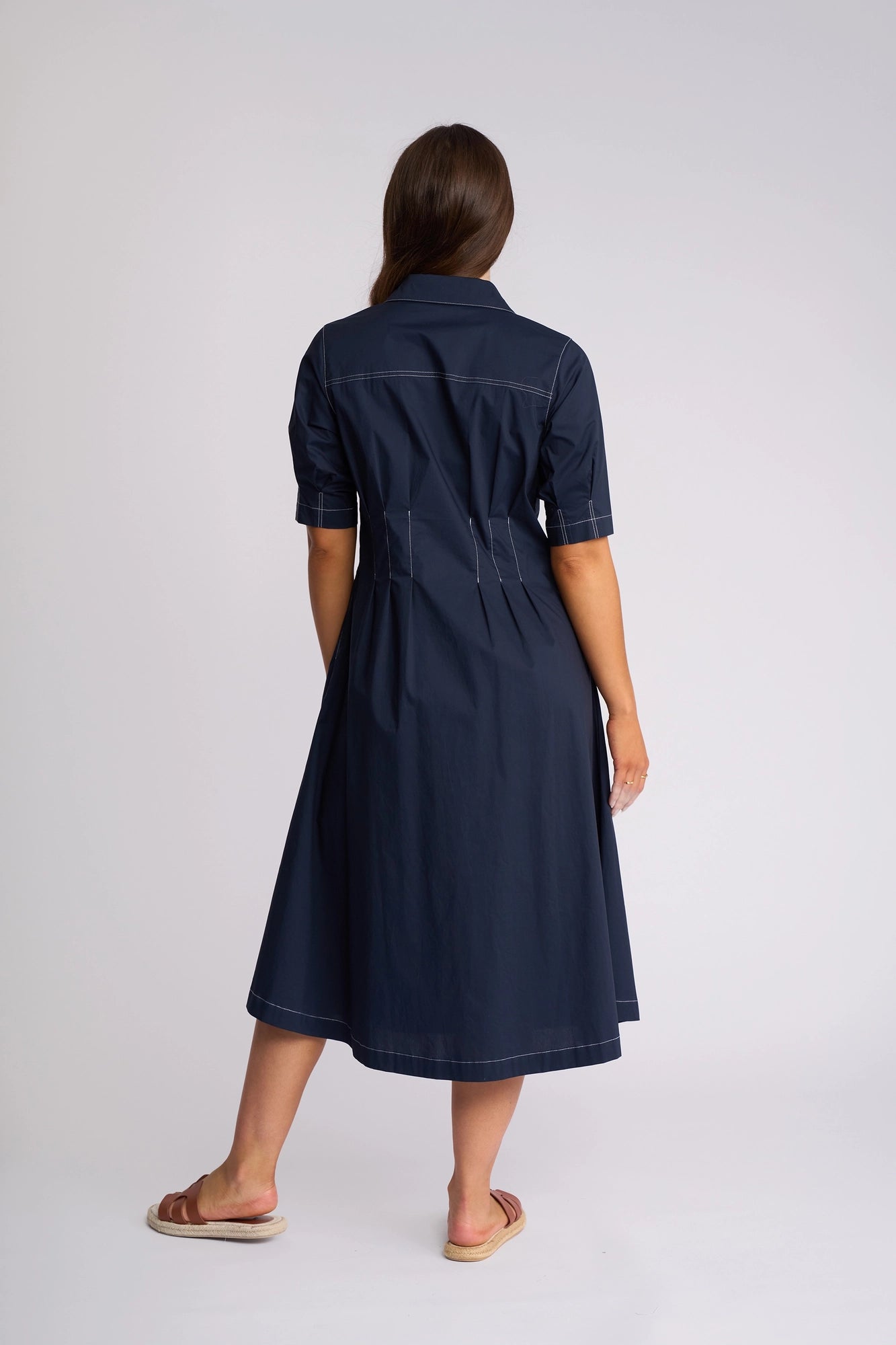 A woman stands facing away, wearing the Vassalli Addie Dress in Ink—a short-sleeve, collared cotton dress featuring white stitching, paired with tan sandals. The feminine silhouette is accentuated by a plain, light-colored background.