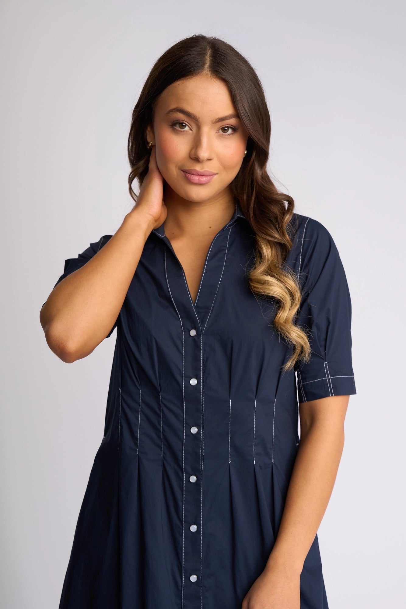 A woman with long brown hair stands against a light background, gently touching her neck and gazing at the camera. She wears the Vassalli Addie Dress - Ink, its feminine silhouette enhancing her graceful presence.