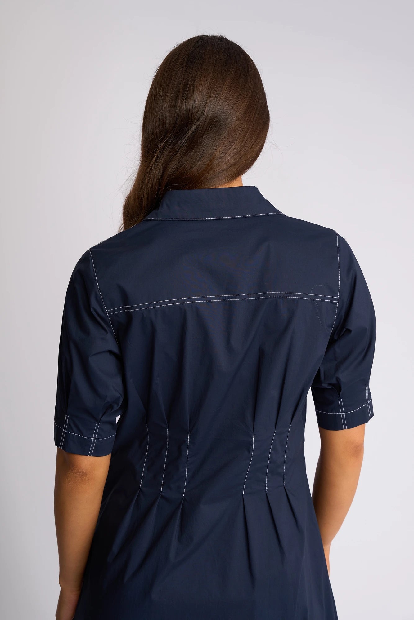 A person with long brown hair is shown from the back, wearing the Vassalli Addie Dress – Ink, a navy blue collared cotton dress with white stitching and short sleeves that beautifully accentuates a feminine silhouette.