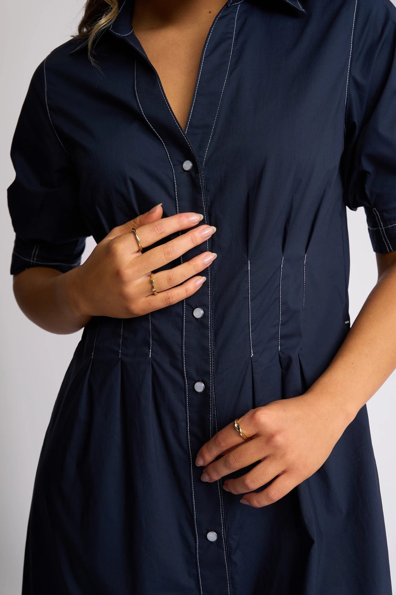A person wearing the Vassalli Addie Dress - Ink stands against a plain background, displaying neatly manicured nails and several gold rings on their fingers. The cotton dress showcases a feminine silhouette with white stitching, merging classic style and modern elegance.