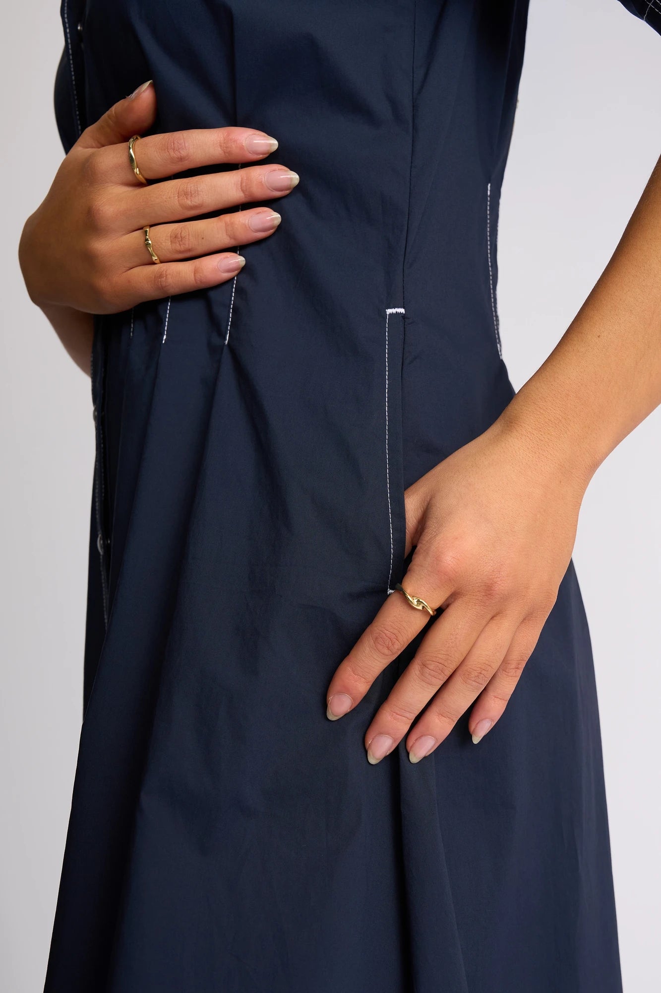 A person wearing the Vassalli Addie Dress - Ink, made from navy blue cotton with white stitching, is shown from the torso down with neatly manicured nails and gold rings enhancing the feminine silhouette of the dress.