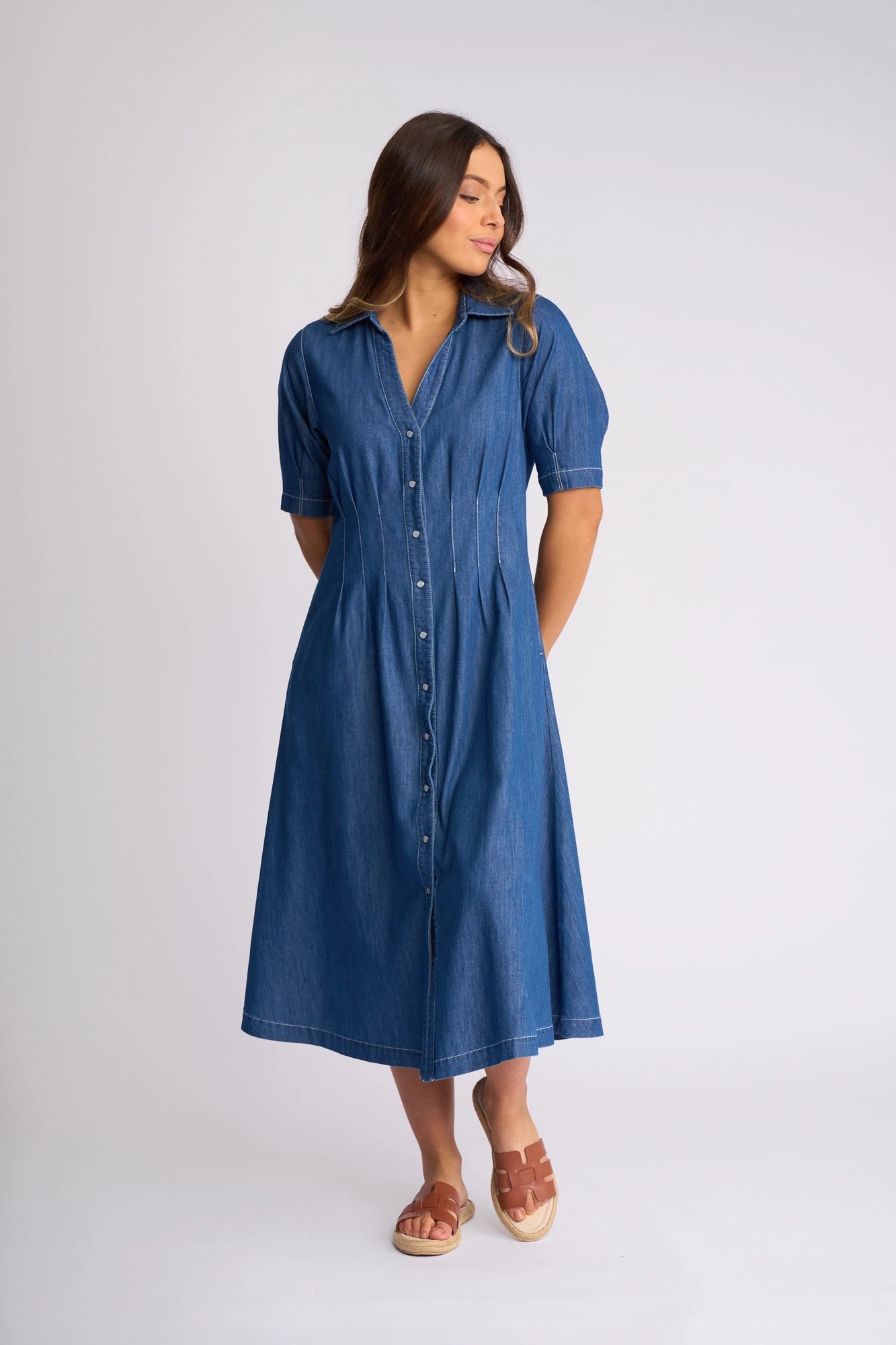 A woman stands against a plain light background, hands behind her back and looking to her left, wearing the Vassalli Addie Dress - Ocean. This blue denim midi dress features short sleeves, a collar, shaped waist, and a classic button-down front with snap fastening. She pairs the dress perfectly with brown sandals.
