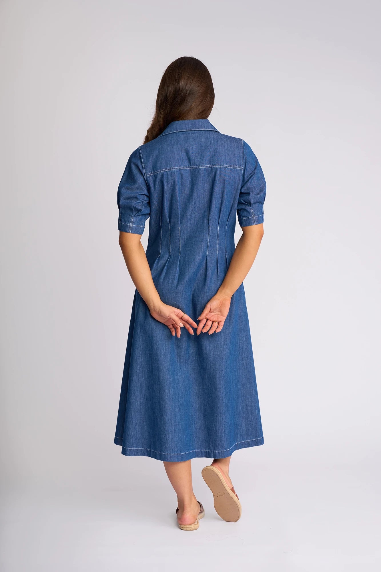 A woman with long brown hair stands barefoot, facing away with her hands behind her back against a plain light background, wearing the Vassalli Addie Dress in Ocean—a blue denim midi dress featuring short sleeves, snap fastening, and a shaped waist.