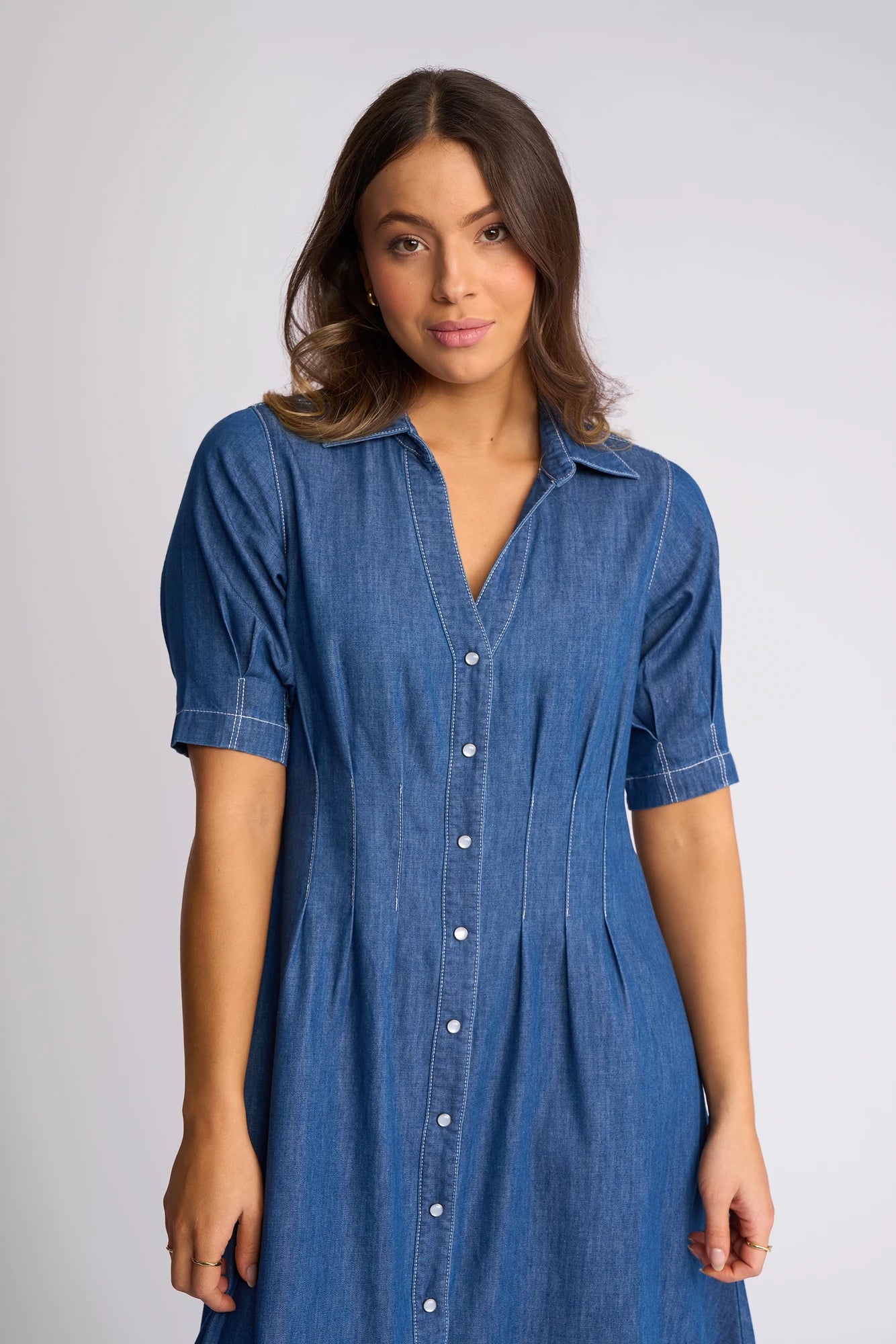 A woman wearing the Vassalli Addie Dress in Ocean—a short-sleeved, button-down blue denim dress featuring snap fastenings and contrast white stitching—stands against a plain light background, looking at the camera with a neutral expression. The dress’s shaped waist subtly accentuates her silhouette.