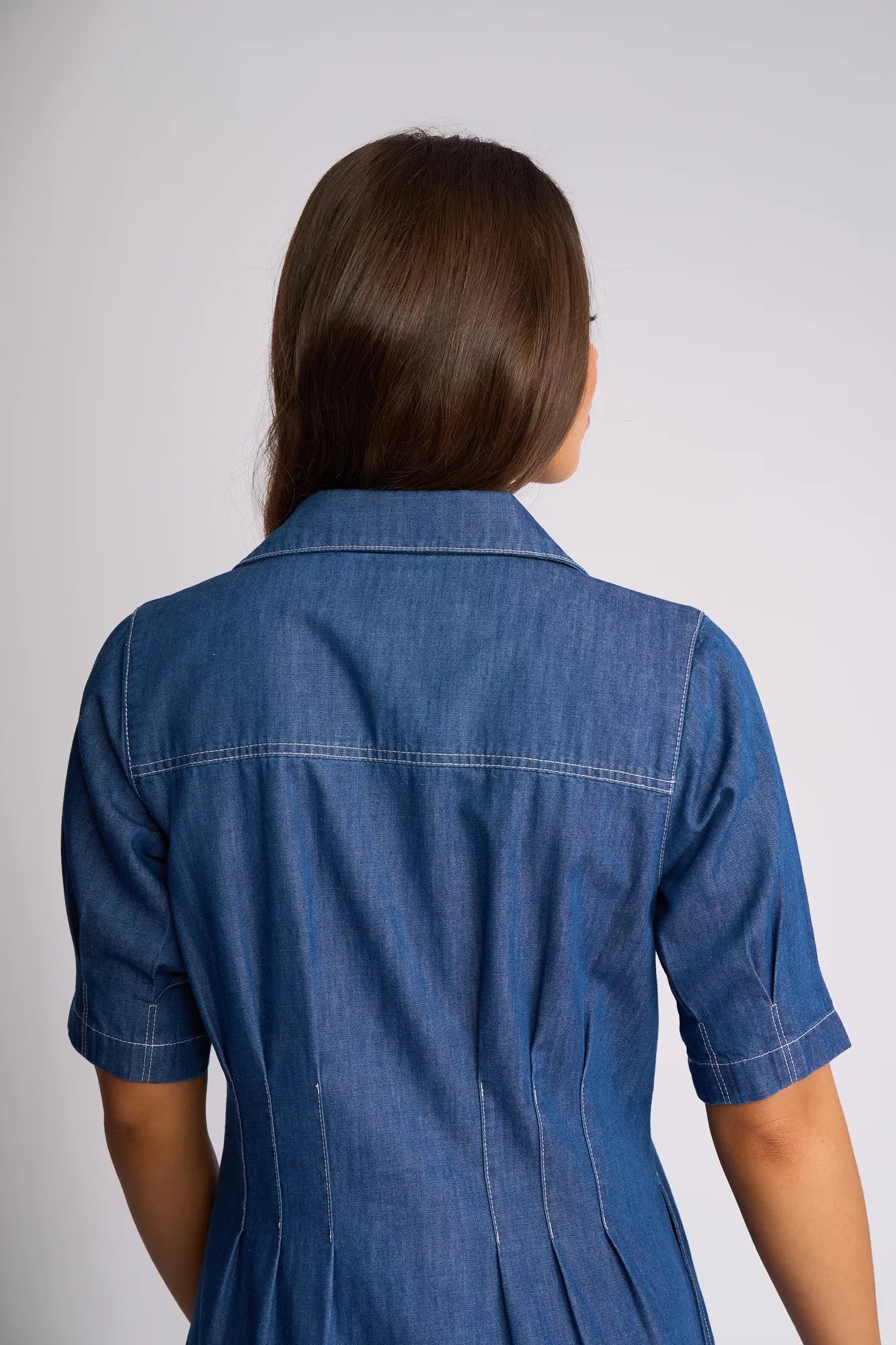 A woman with long brown hair, seen from behind, is wearing the Vassalli Addie Dress in Ocean, a short-sleeve blue denim dress featuring a shaped waist and white stitching.