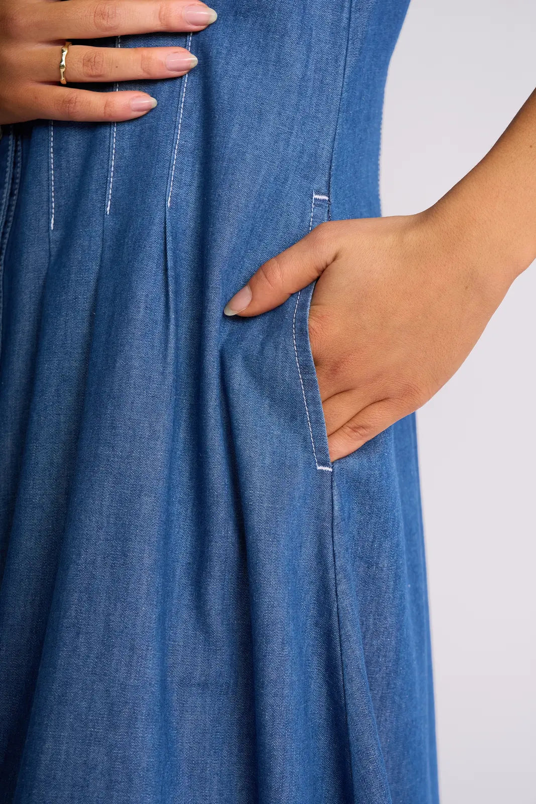 A person wearing the Vassalli Addie Dress - Ocean, a blue denim dress with a shaped waist, places one hand in the pocket and rests the other on their waist. The dress features white stitching, a flared skirt, and snap fastenings down the front.