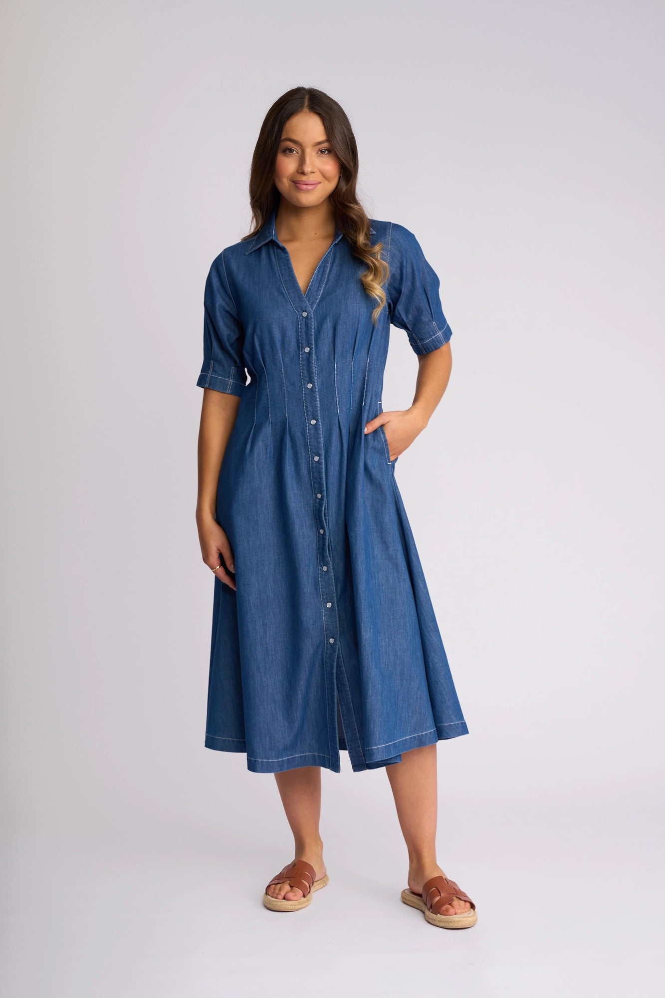 A woman stands against a light background, smiling with one hand in her pocket, wearing the Vassalli Addie Dress in Ocean—a blue, button-down, knee-length denim dress featuring short sleeves and a shaped waist—paired with brown sandals. Her long hair cascades over her shoulders.