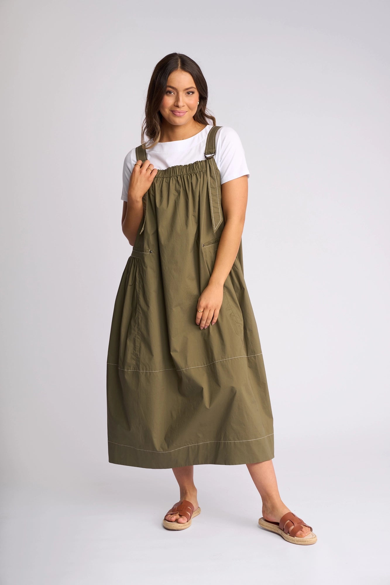 A woman stands in a studio wearing the Vassalli Blend Dress in Khaki, layered over a white t-shirt. Featuring shoulder ties and a loose cotton fit, she pairs the look with sandals and has medium-length hair, smiling softly while holding one of the dress straps.
