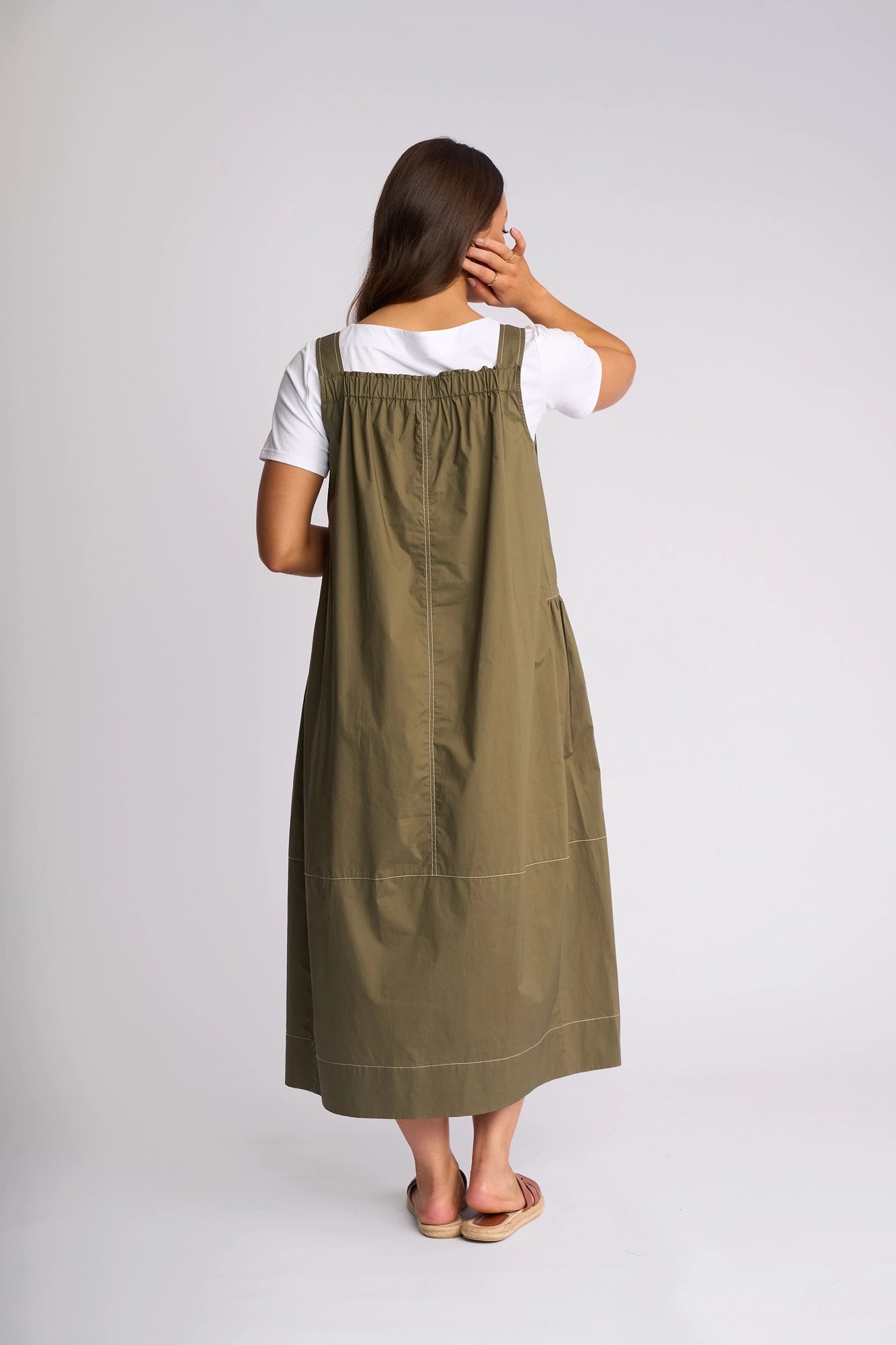 A woman stands facing away, wearing the Vassalli Blend Dress in khaki—a long cotton pinafore style layered over a white t-shirt. Her long brown hair falls down her back as she touches her face with one hand, sandals completing the effortless summer look against a plain white background.