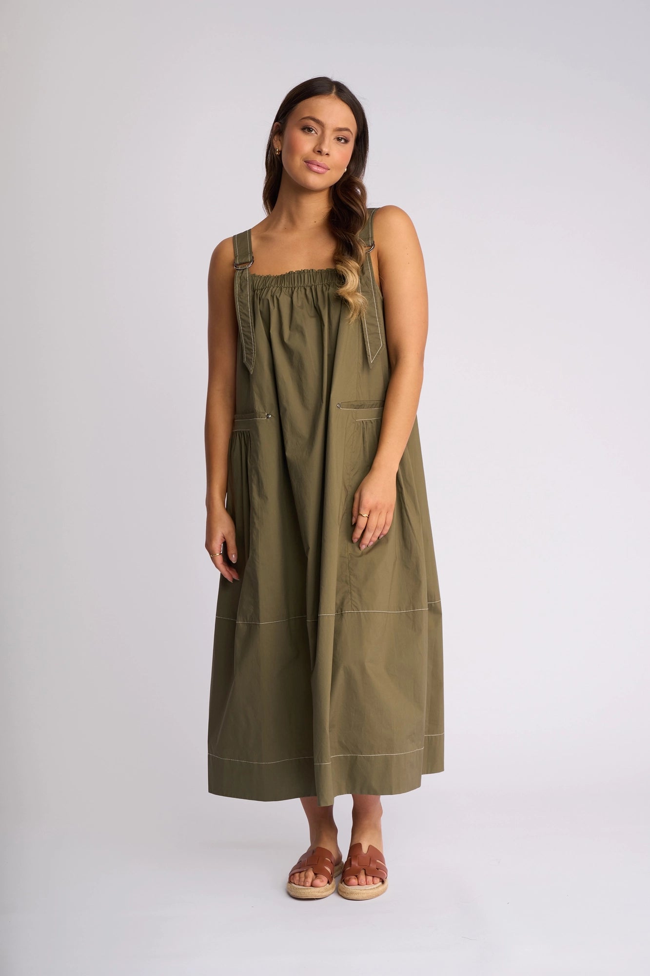 A woman stands against a plain backdrop wearing the Vassalli Blend Dress in Khaki, a sleeveless maxi ideal for summer, paired with brown sandals. Her hair cascades in loose waves over one shoulder.