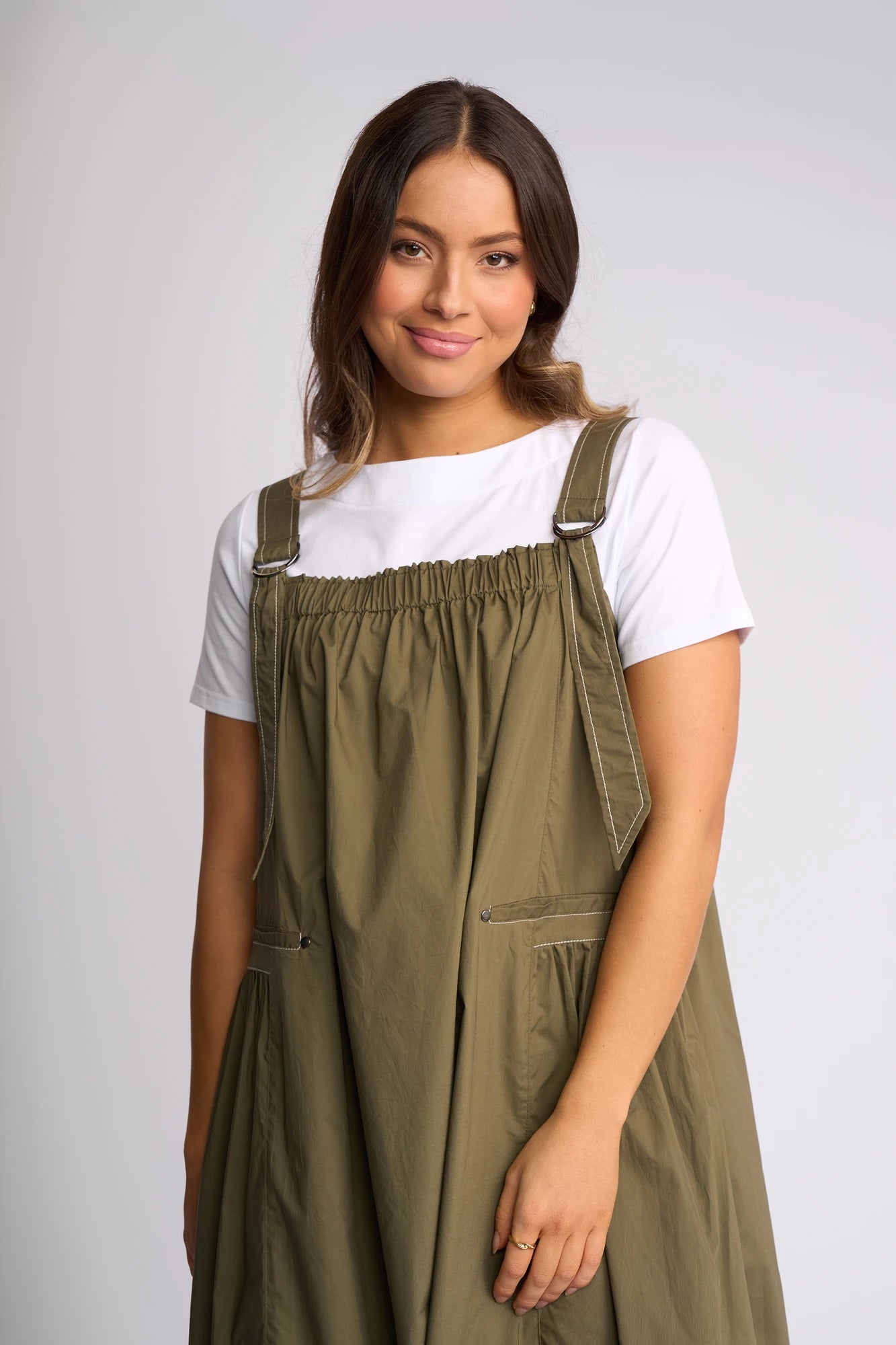 A woman with long brown hair wears the Vassalli Blend Dress in khaki over a white t-shirt, standing against a plain light background and smiling gently at the camera.
