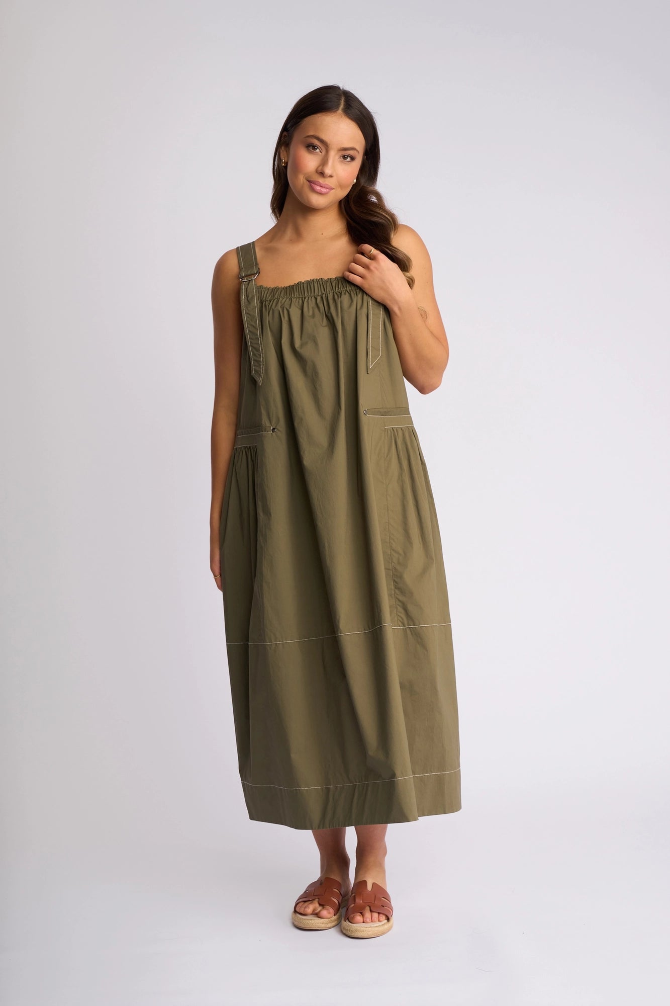 A woman stands against a plain backdrop, wearing the Vassalli Blend Dress in khaki—a sleeveless, ankle-length summer dress with a gathered top—paired with brown sandals. Her long brown hair and relaxed expression capture the effortless style of this Vassalli piece.