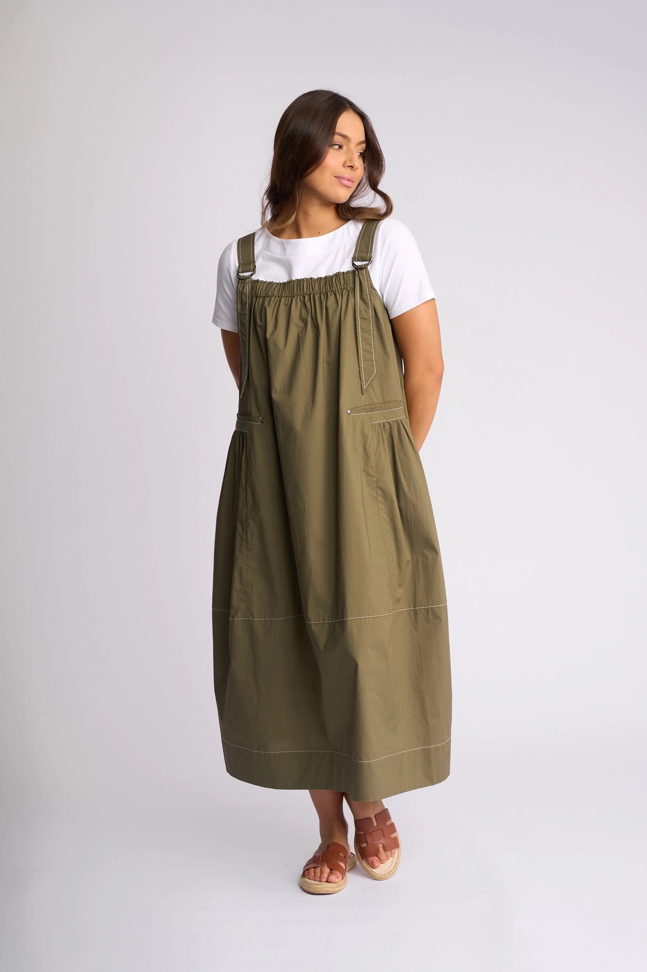 A woman stands against a plain light background, wearing the Vassalli Blend Dress in Khaki—an olive-green cotton pinafore style with large pockets—layered over a white short-sleeve shirt and paired with brown sandals.