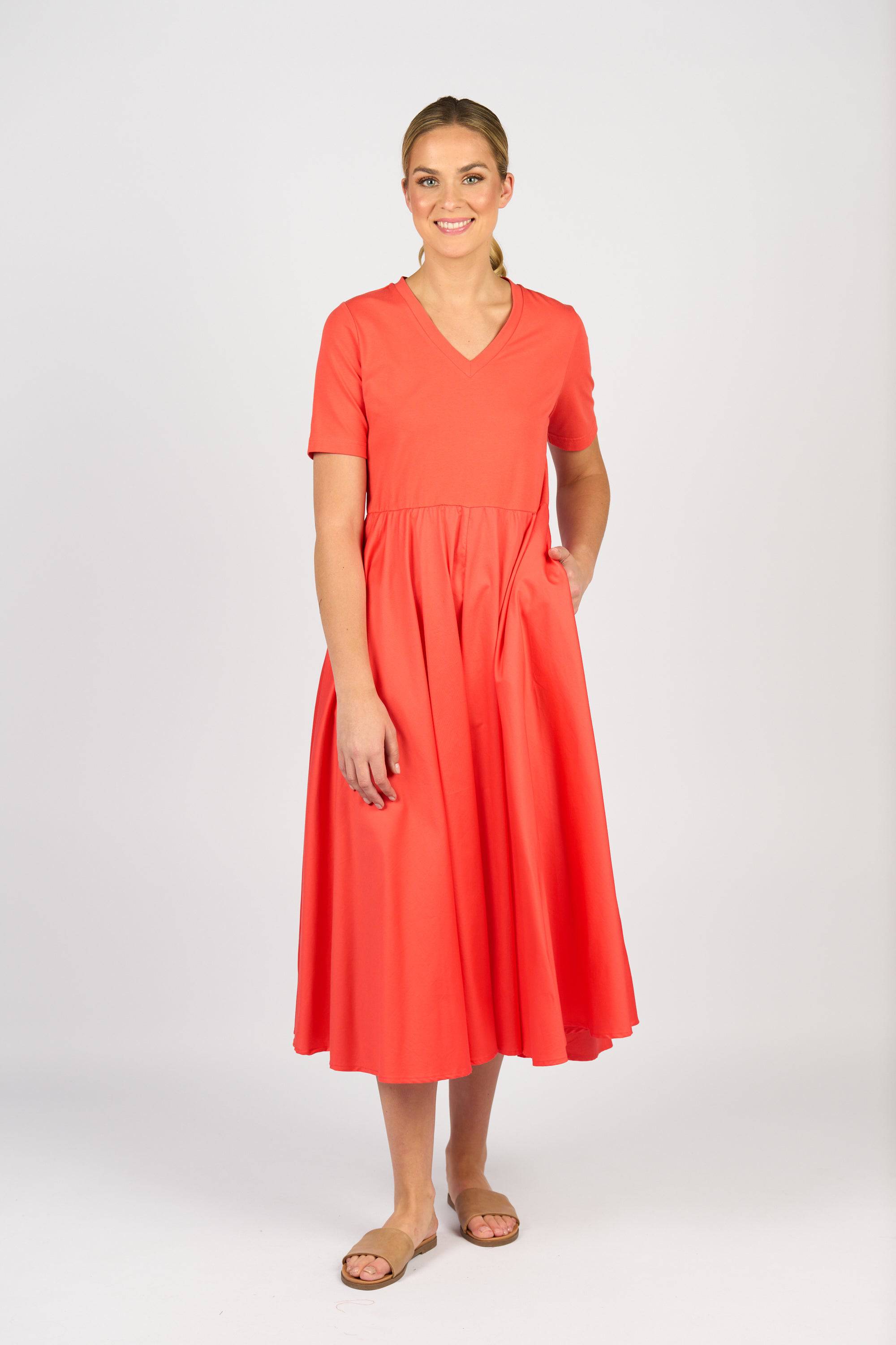 A woman smiles in the Vassalli T-Shirt Dress - Aperol, featuring a coral hue, V-neck, short sleeves, fitted waist, and flared skirt. She pairs it with tan flat sandals and stands against a plain white background with her hair pulled back.