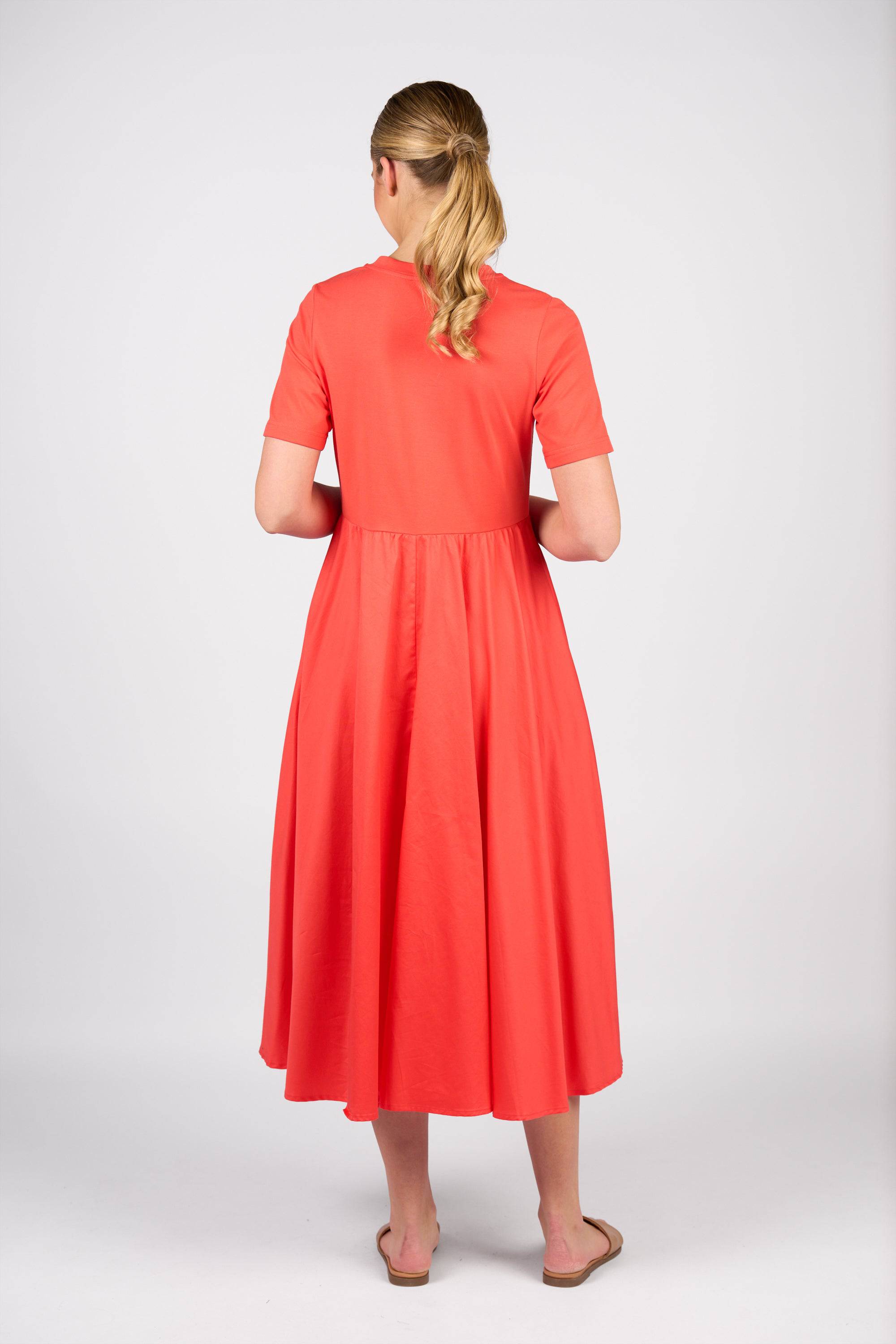 A woman with blonde hair in a low ponytail is barefoot and facing away from the camera, wearing the Vassalli T-Shirt Dress - Aperol against a plain light background.