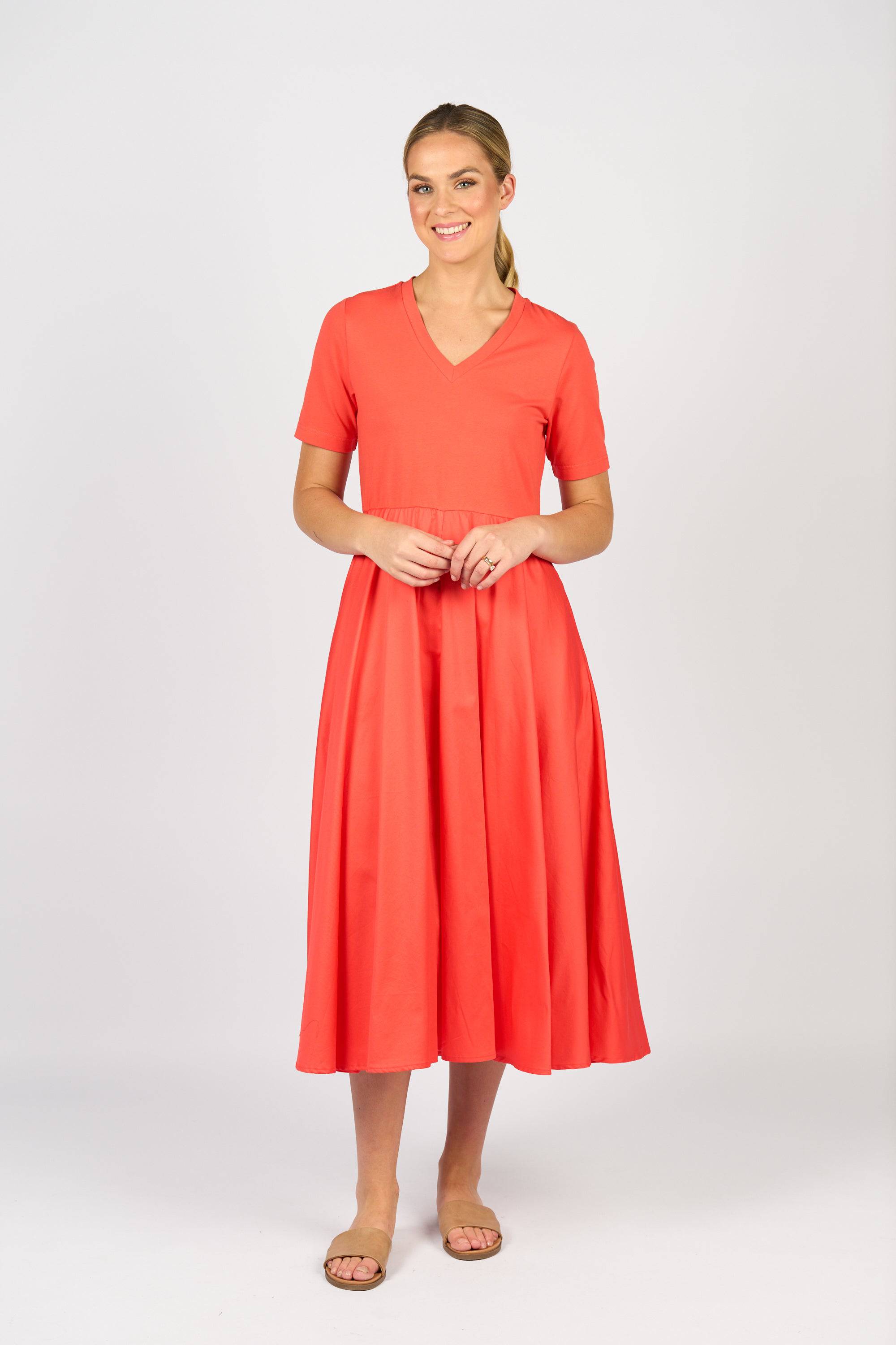 A woman smiles in the Vassalli T-Shirt Dress - Aperol, featuring a vibrant coral hue, soft cotton-elastane blend, fitted waist, and flared skirt. She pairs it with flat sandals and poses with her hair tied back against a light background.