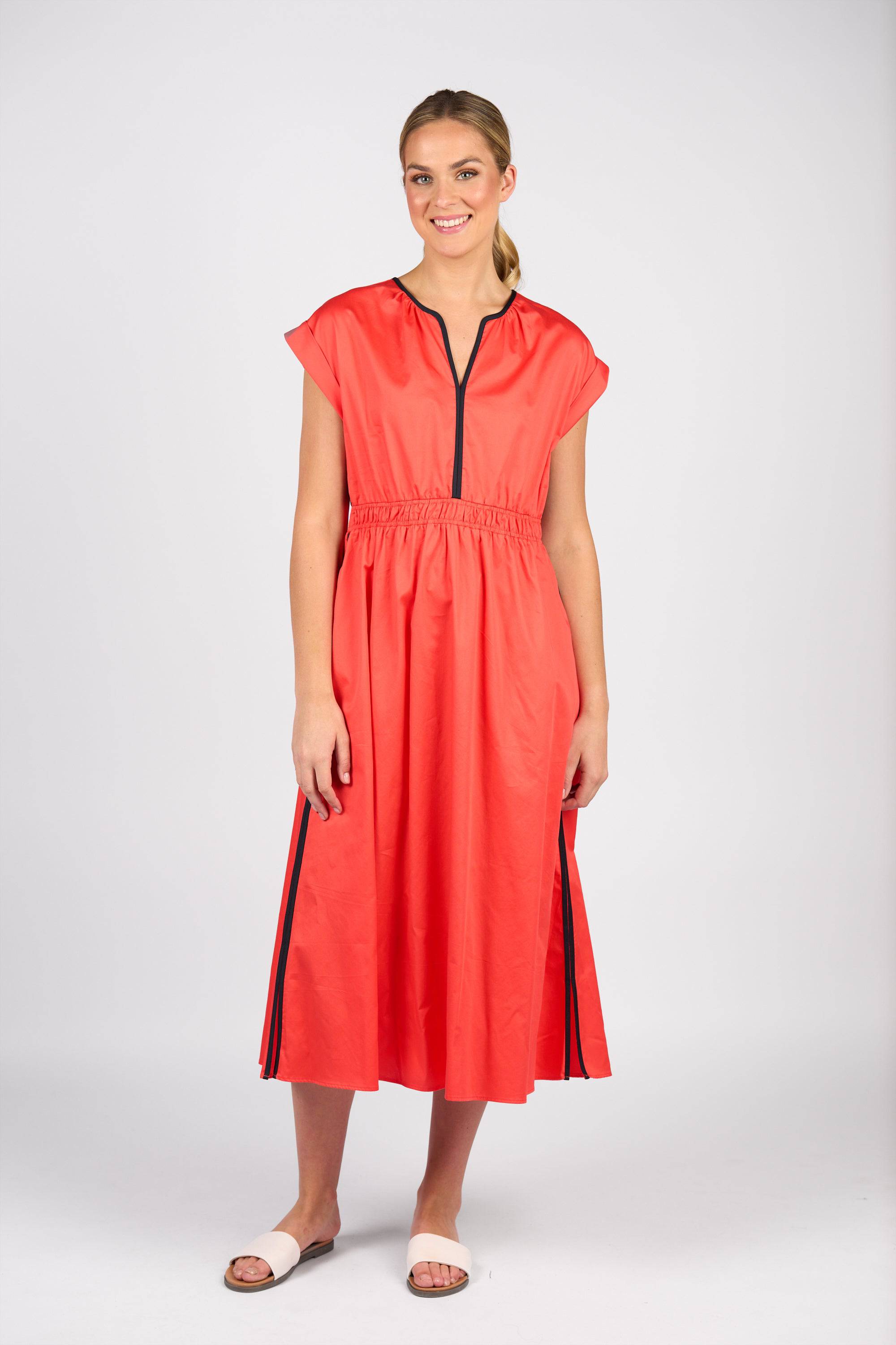 A woman in a ponytail and white sandals wears the Vassalli Emmie Dress - Aperol/Ink, a coral-red short-sleeved cotton dress with black stripes down the sides, v-neckline, gathered elastic waist, and black zipper detail against a plain background.