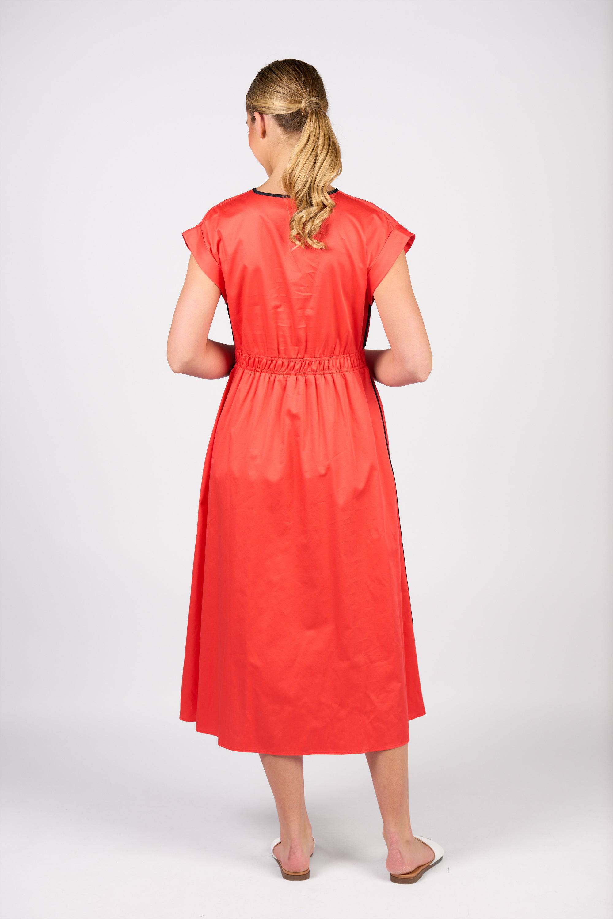 A woman with blonde hair in a ponytail stands facing away, wearing the Vassalli Emmie Dress in Aperol/Ink against a plain white background.