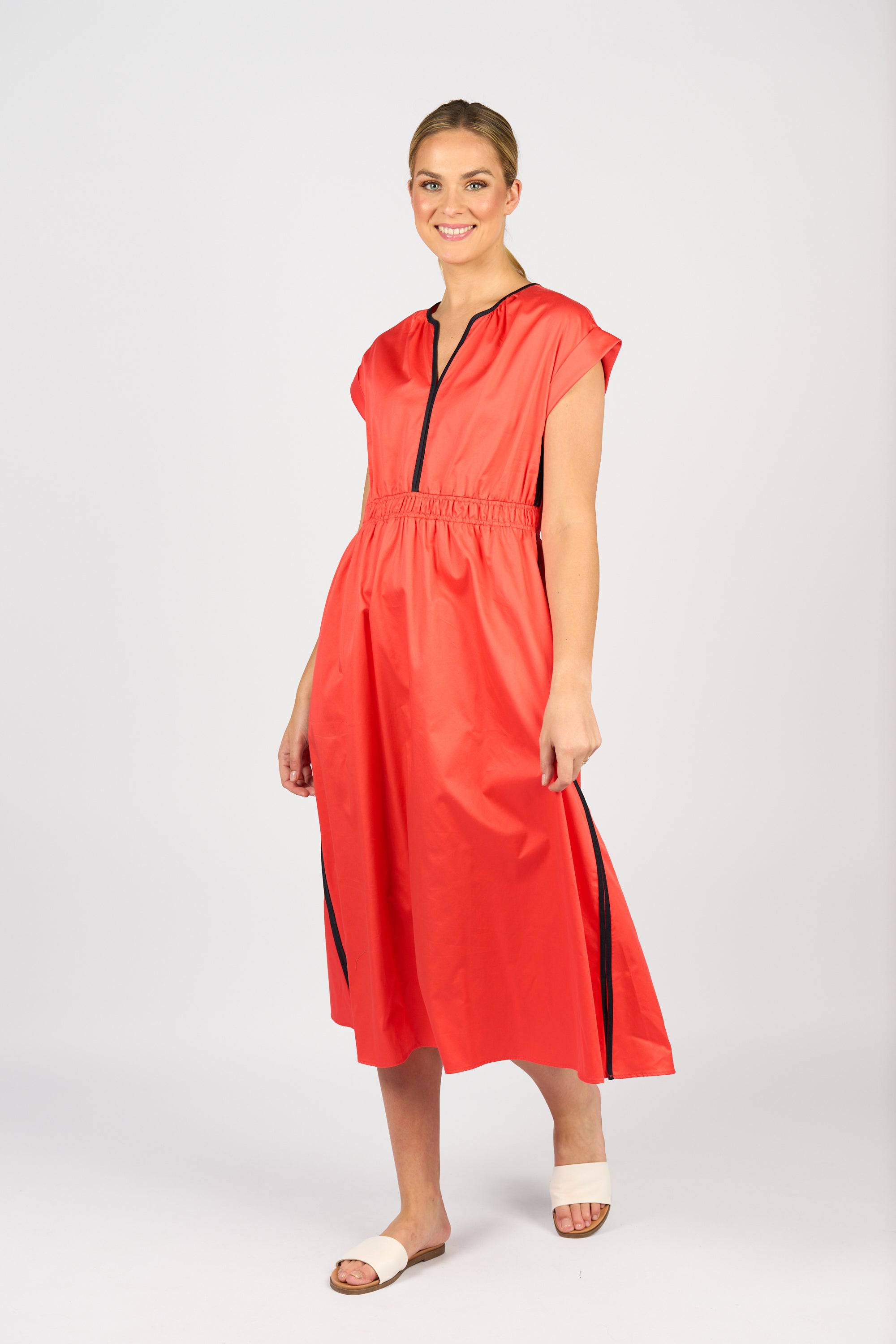 A woman smiles in the Vassalli Emmie Dress - Aperol/Ink, featuring short sleeves, a v-neckline, black zipper and side stripes. She pairs it with white slide sandals and stands against a plain light background.