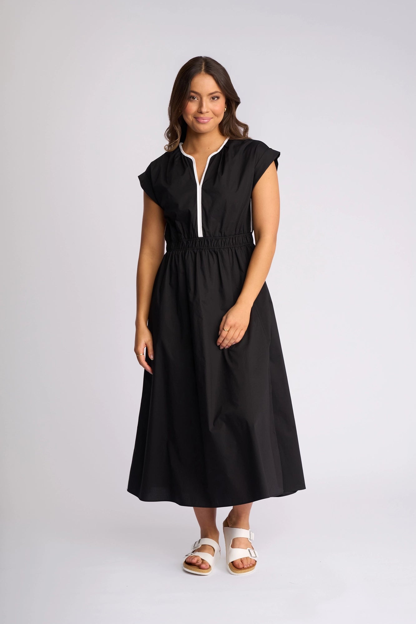 A woman wearing the Vassalli Emmie Dress in Black/White stands against a plain white background. The short-sleeve, cotton midi dress features a relaxed silhouette, breathable fabric, and is accented with a white zipper detail. She has medium-length brown hair and pairs the look with white sandals.