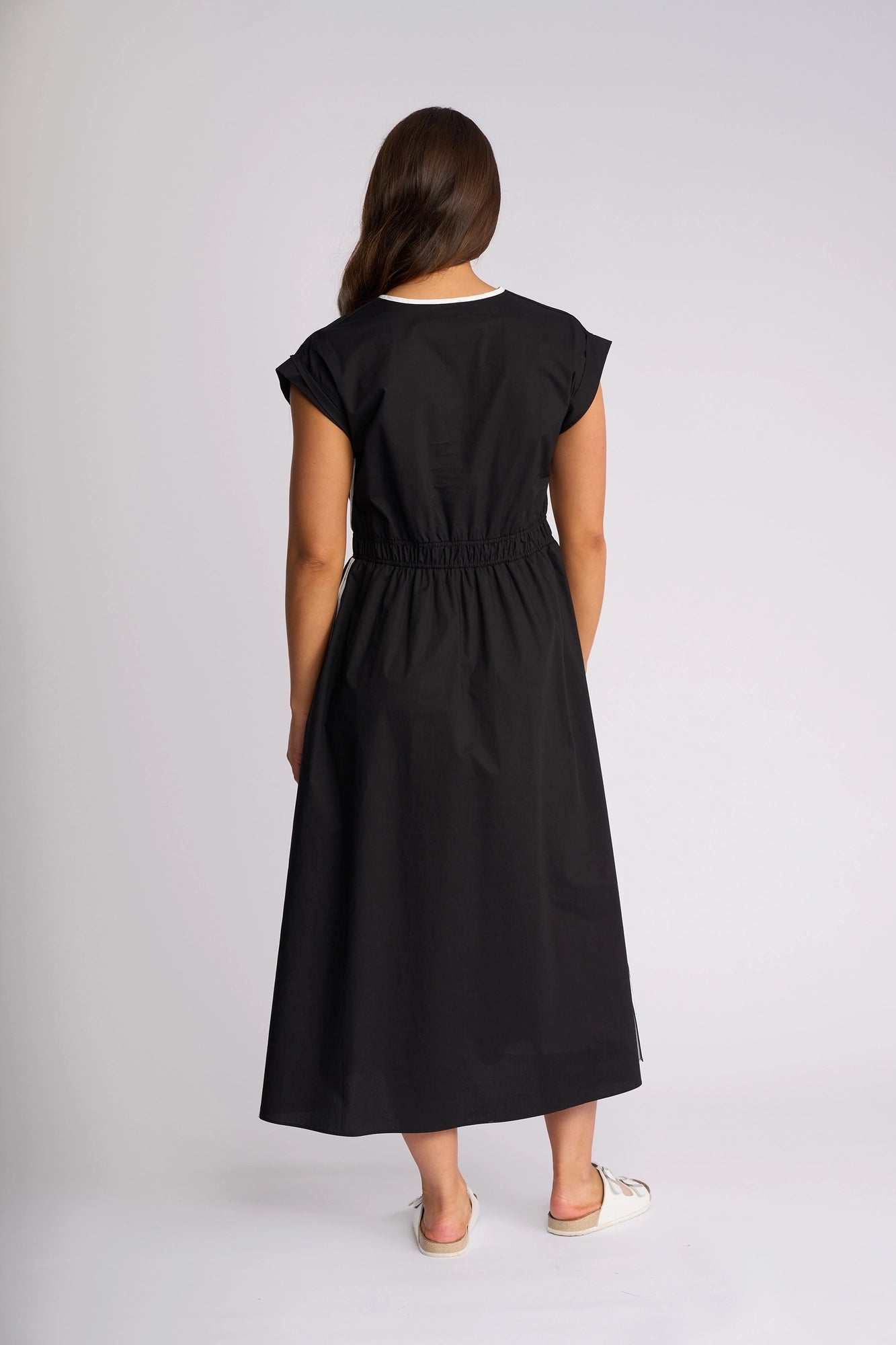 A woman with long brown hair is seen from behind in a studio setting with a plain background, wearing the Vassalli Emmie Dress in Black/White—a relaxed silhouette black cotton midi dress with short sleeves—paired with white slip-on shoes.